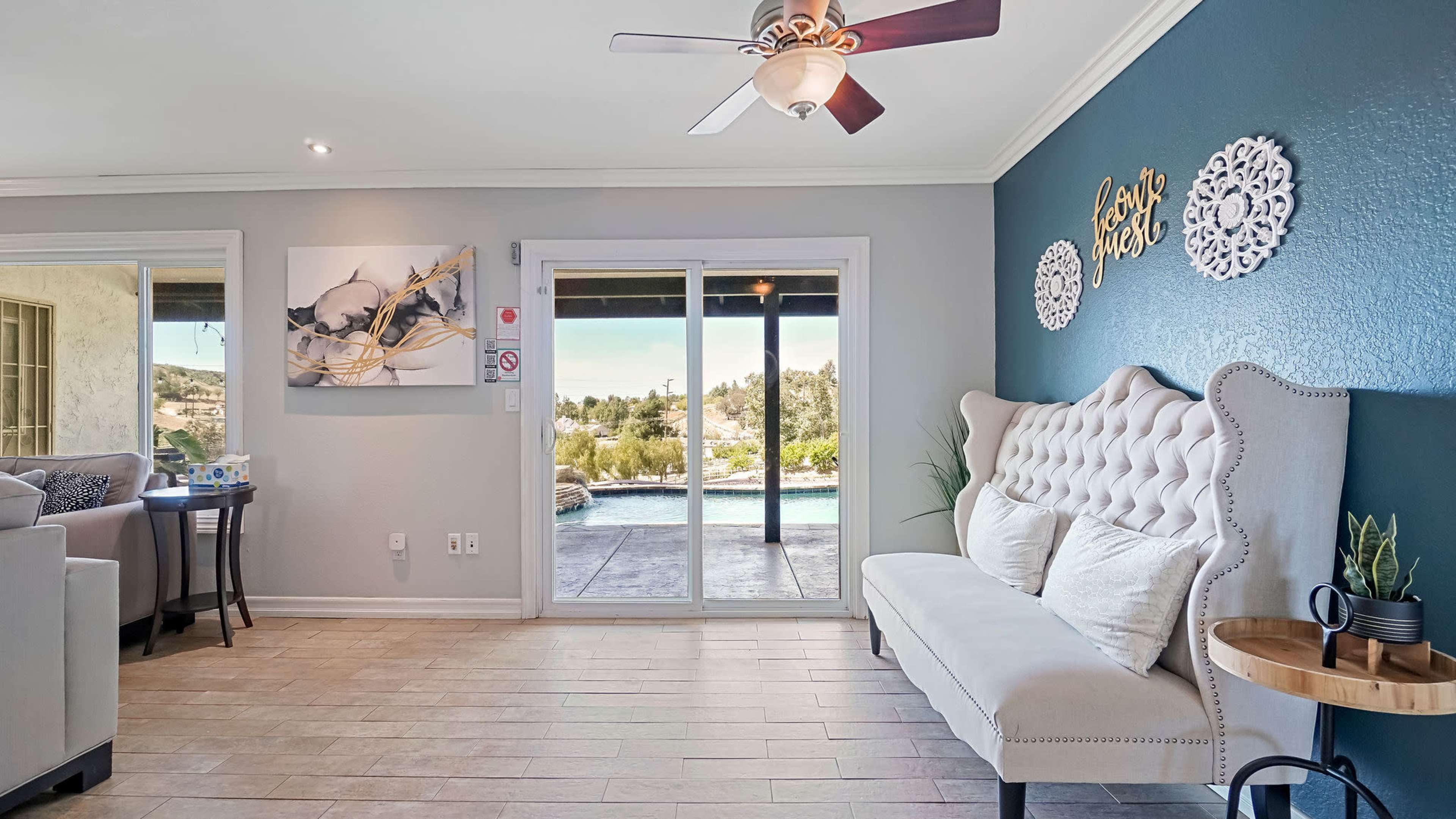 A light-colored living room with a tufted bench against a blue accent wall, leading to large glass doors that open to a view of an outdoor pool area.