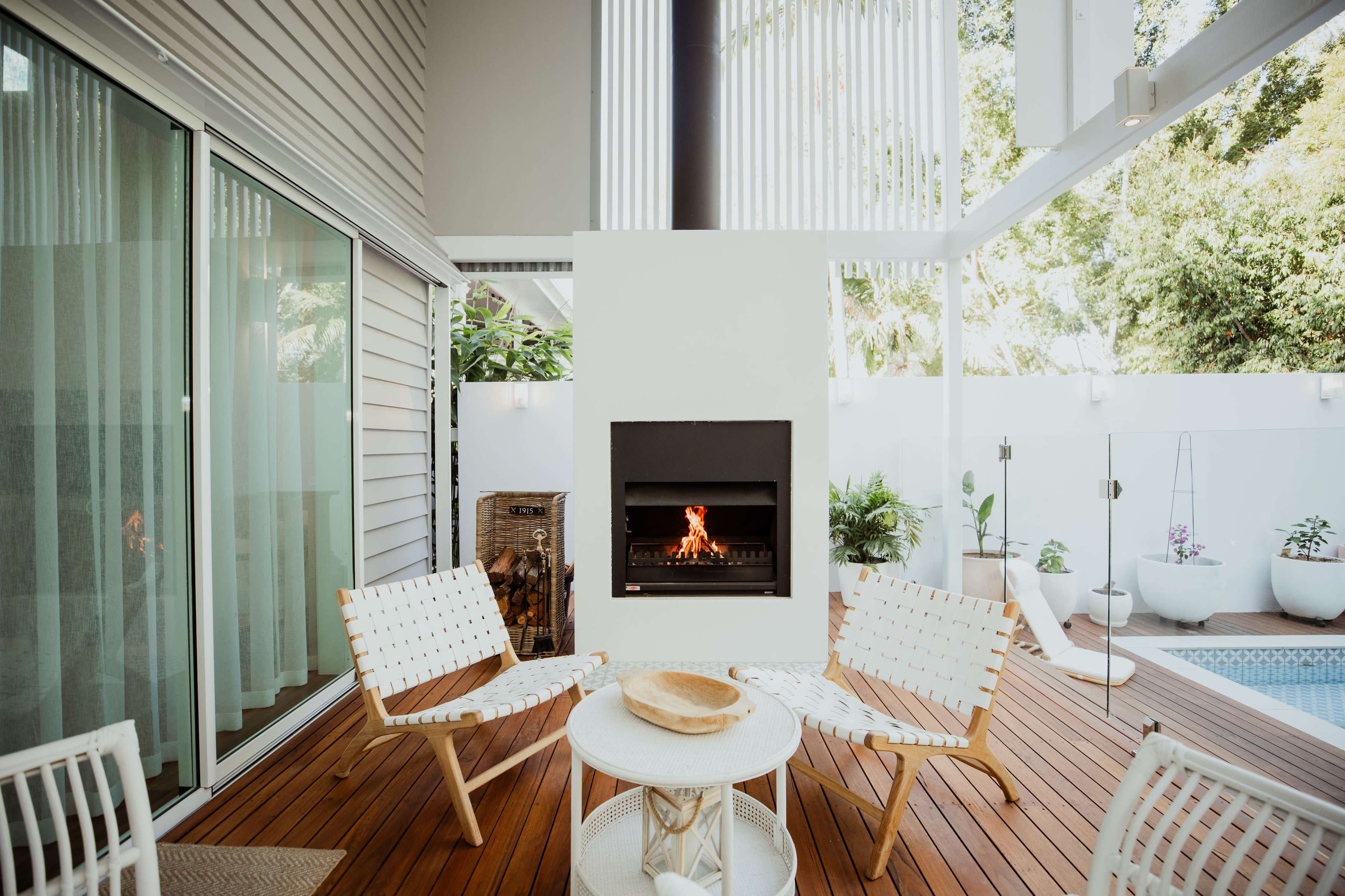 The image shows a modern outdoor living space featuring two wooden chairs, a round table, and a wall-mounted fireplace, surrounded by greenery and a swimming pool.