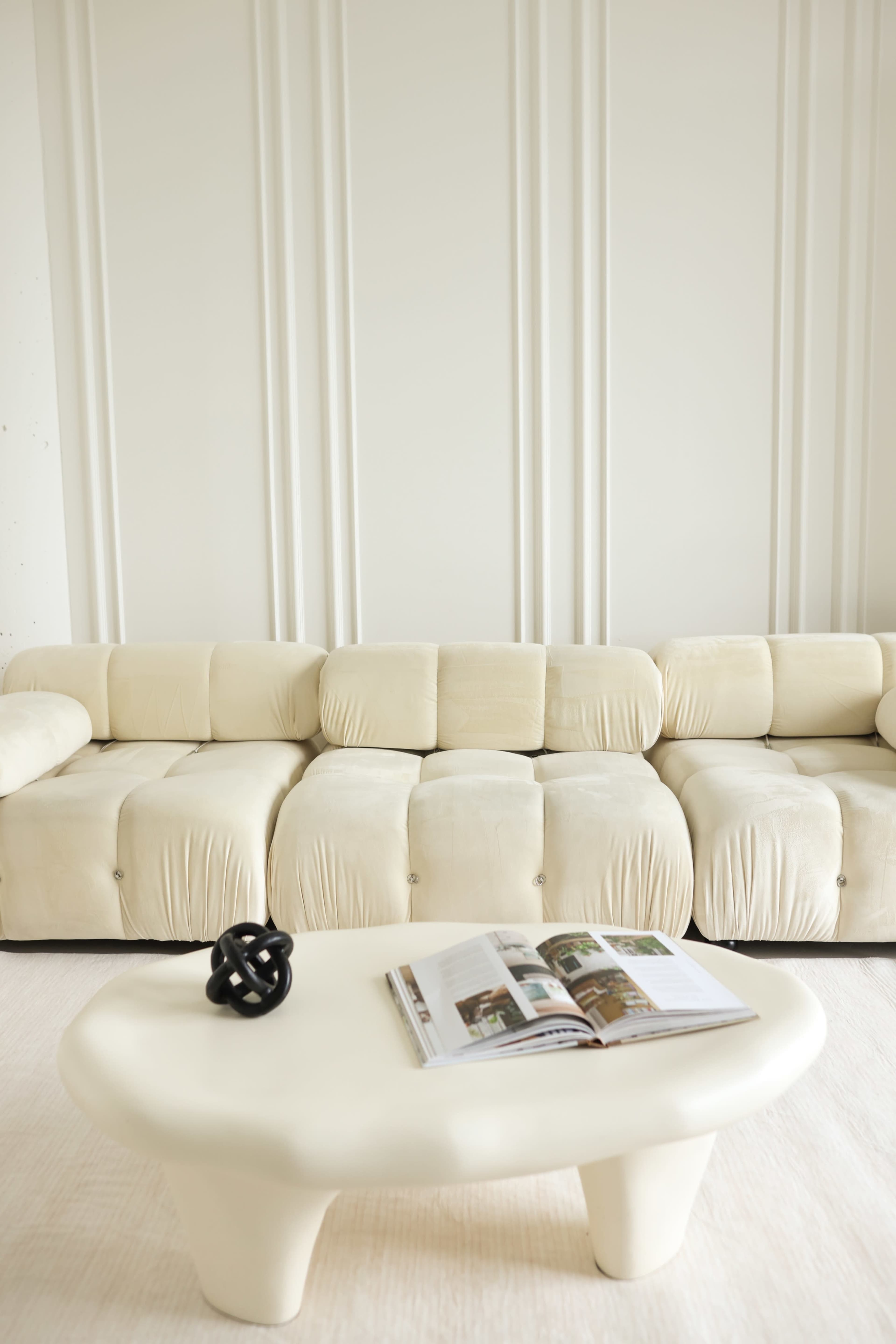 A cream-colored sectional sofa is positioned next to a round coffee table featuring an open magazine atop it, against a plain wall with vertical paneling.