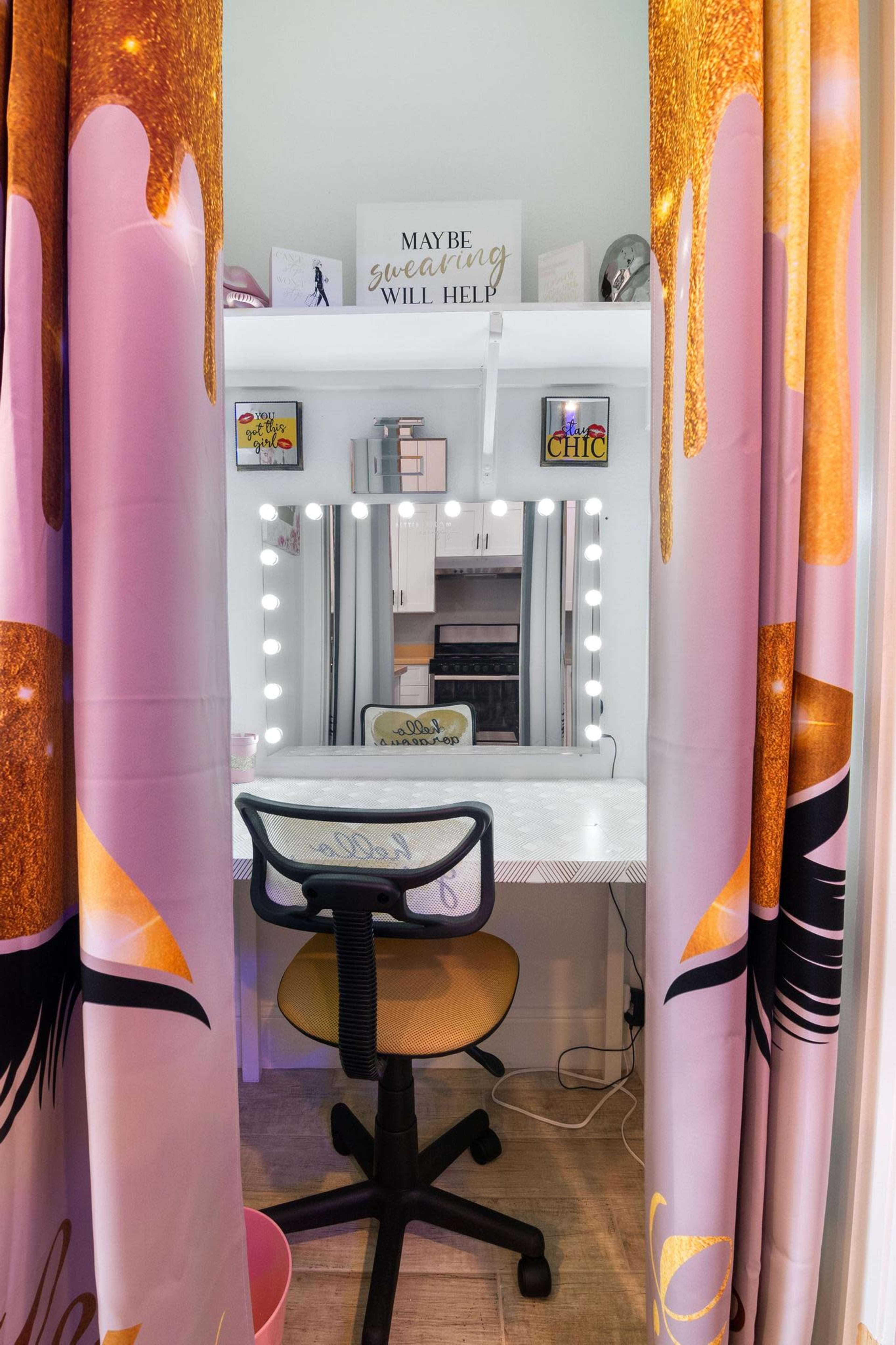 A vanity area is framed by decorative curtains, featuring a mirror with lights and a swivel chair, against a backdrop of a motivational sign.