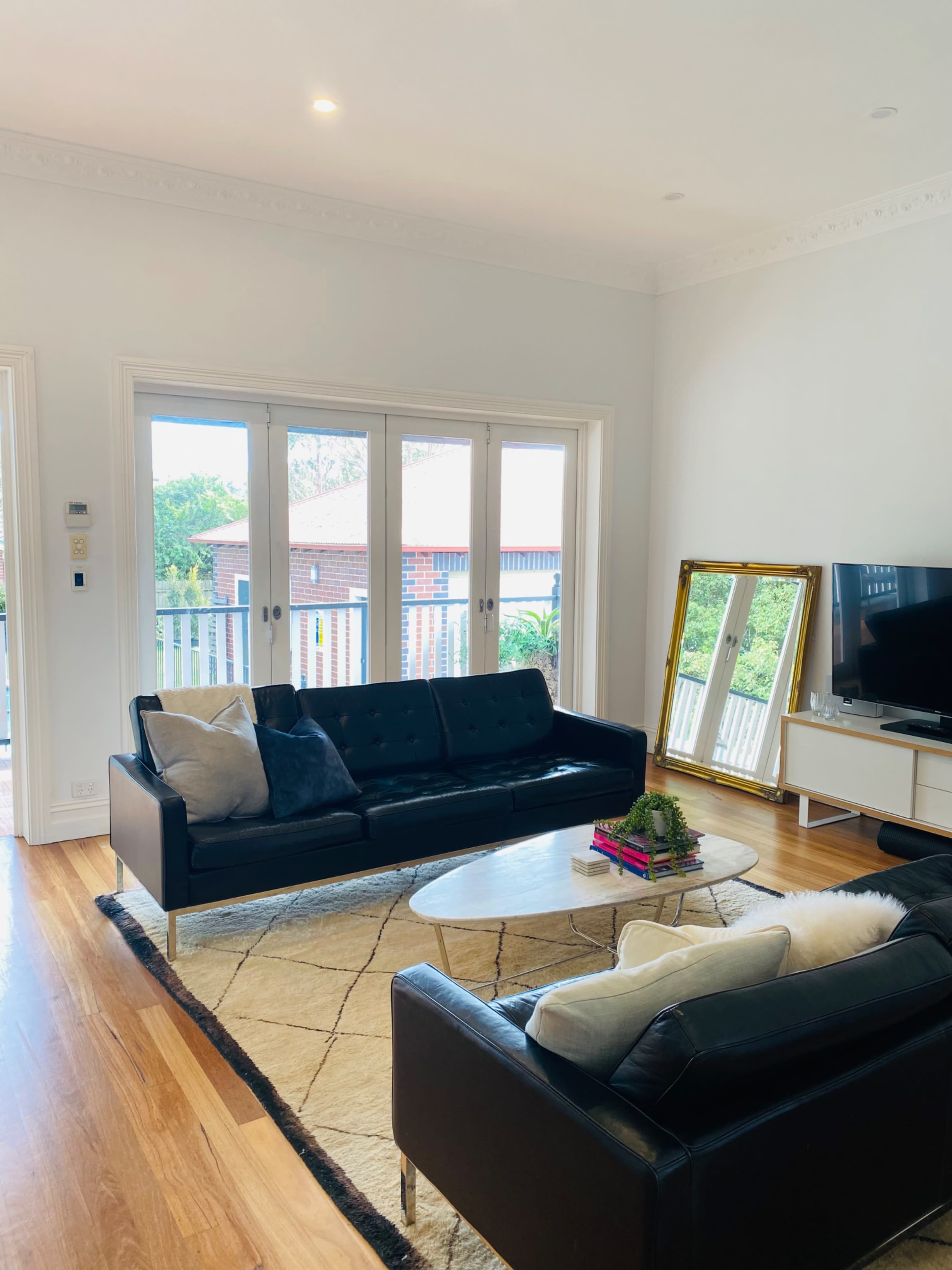 The living room features two black sofas facing a coffee table on a rug, with large glass doors opening to an outdoor area and a mirror leaning against the wall.