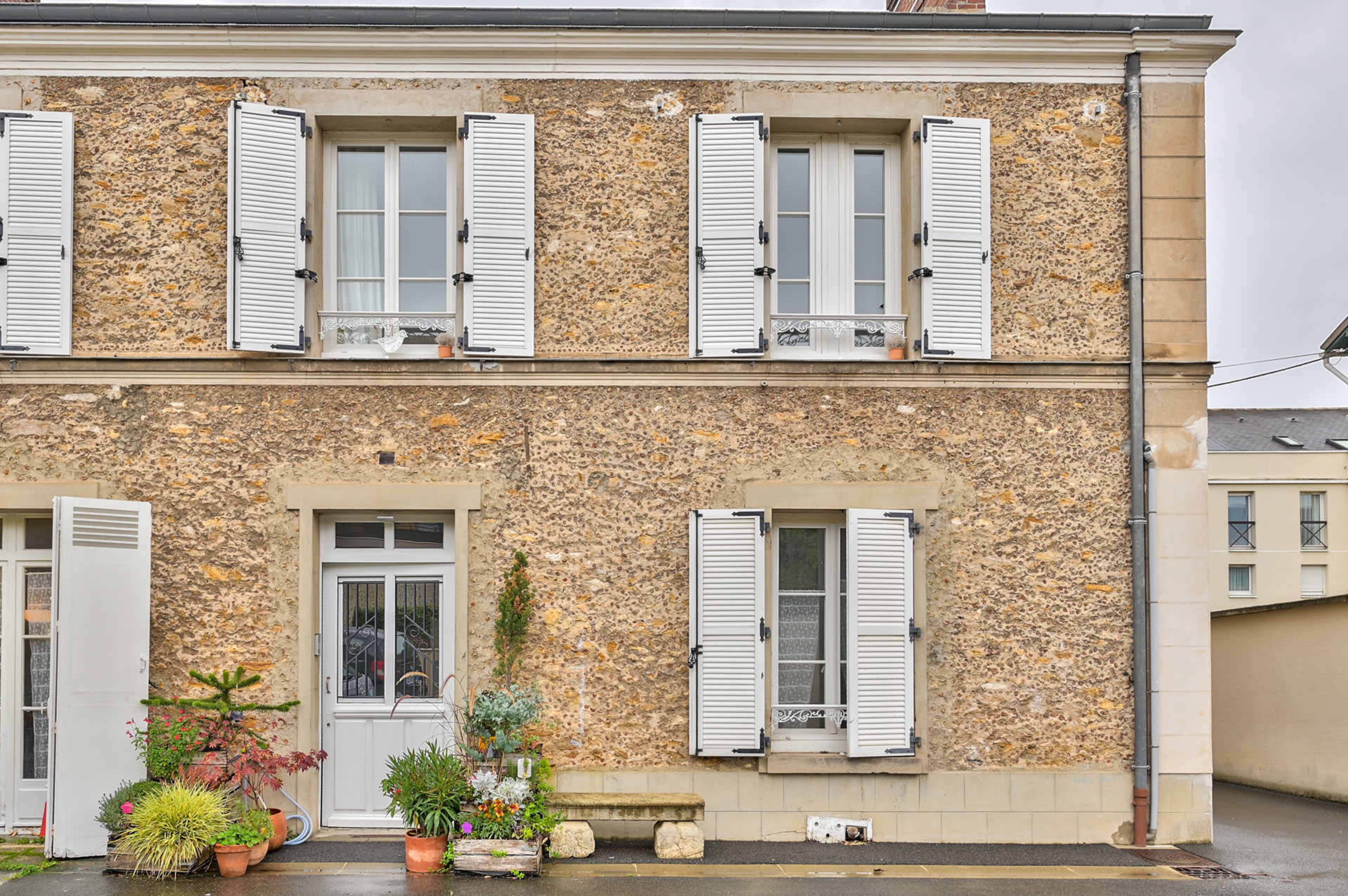 The image shows a two-story house with a textured stone facade, white shutters, and a front door surrounded by potted plants.