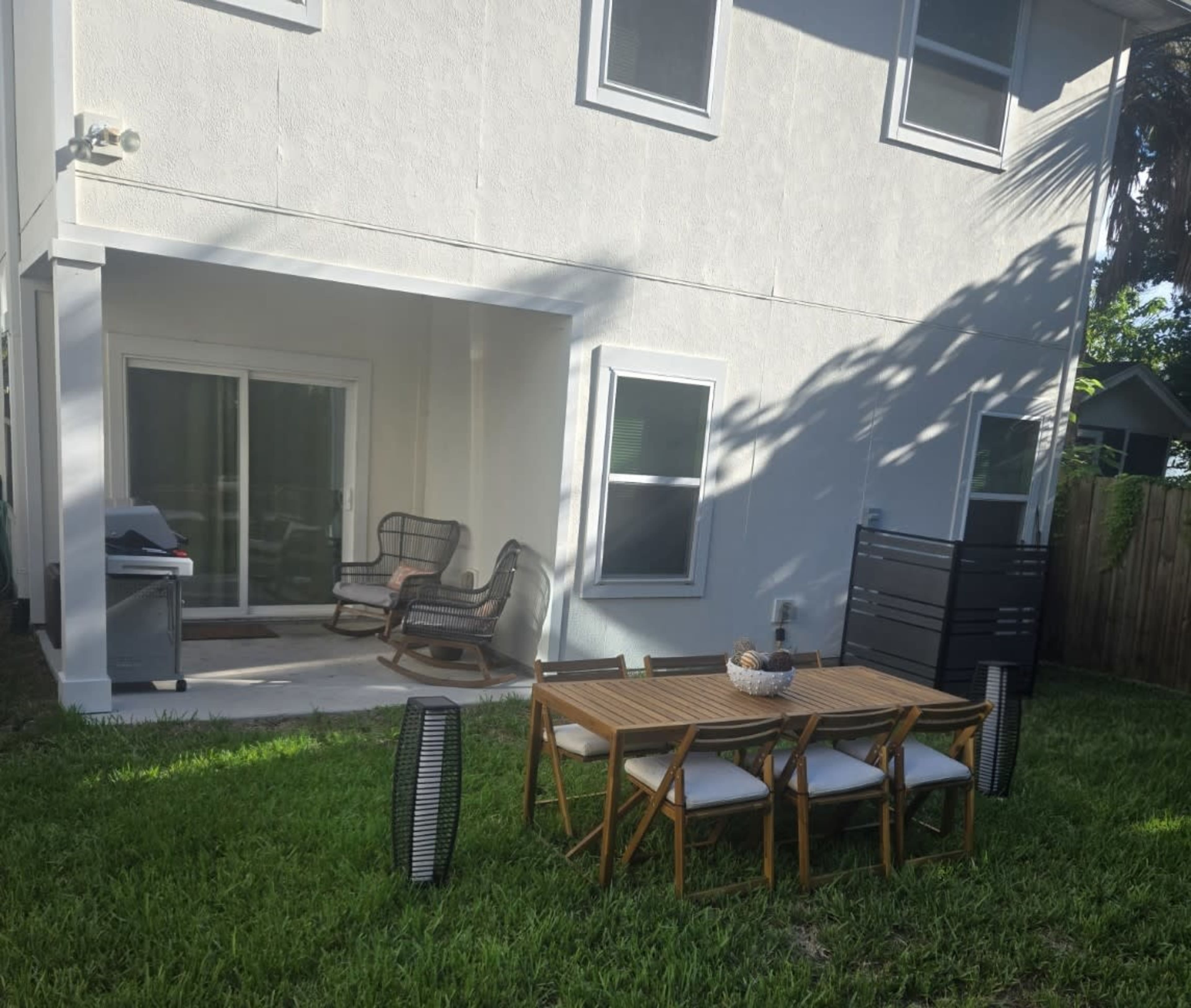 The image shows a backyard with a wooden dining table and chairs on grass, a covered patio with a rocking chair, and a grill.