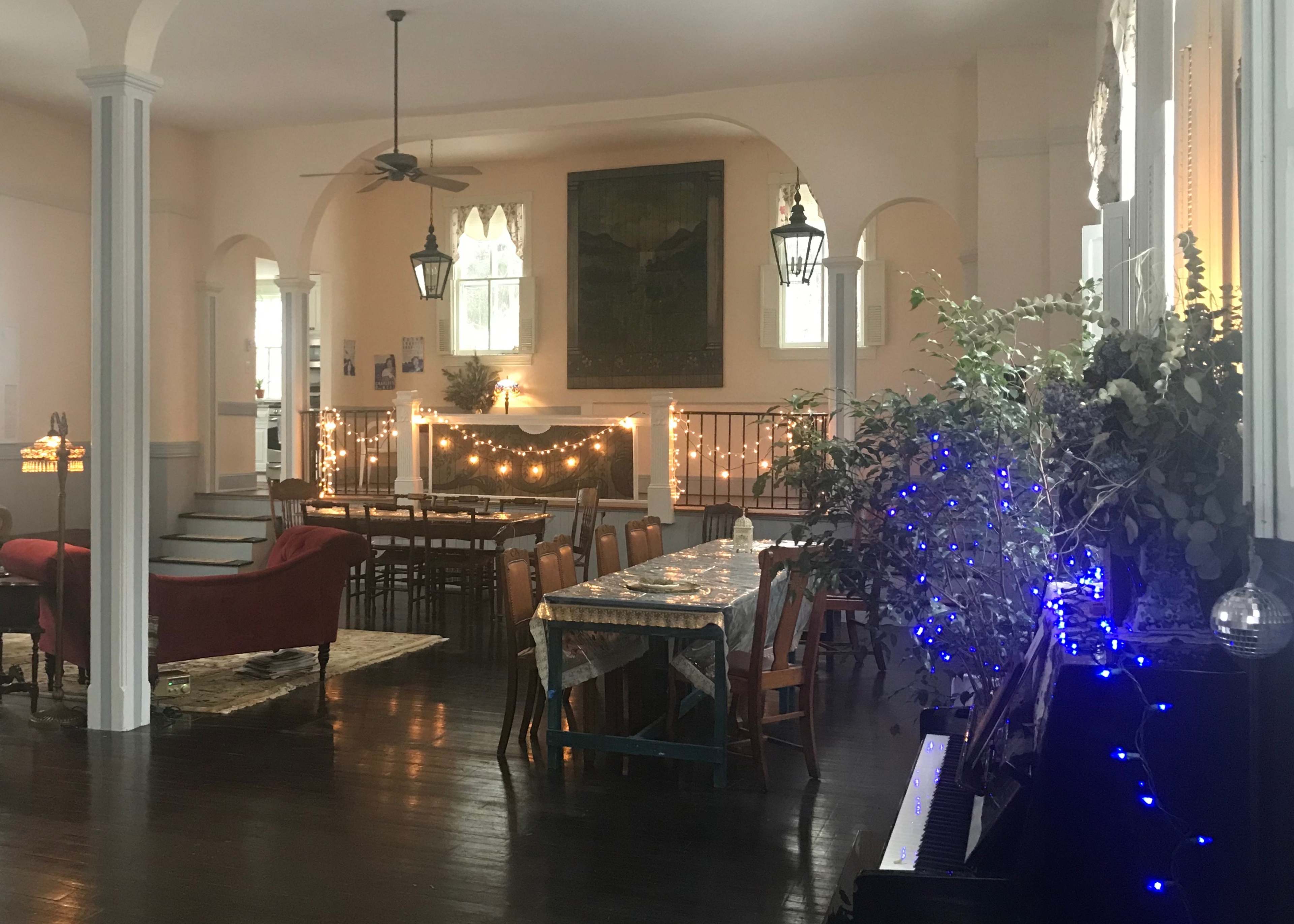A spacious room features a long table surrounded by chairs, with string lights and decorative plants adding ambiance to the setting.