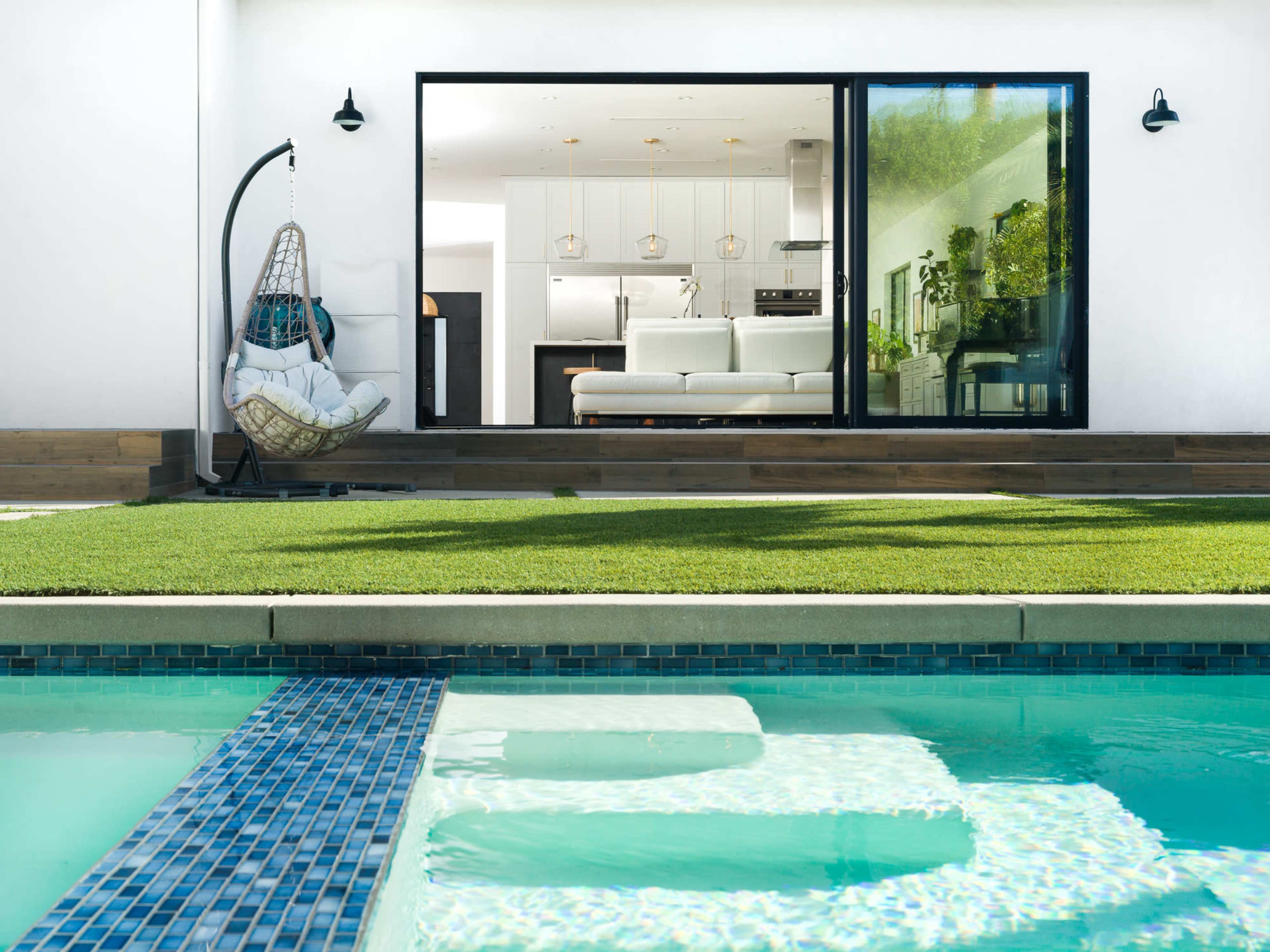 A glass sliding door opens from a modern living space to a backyard with a pool and a neatly manicured lawn.