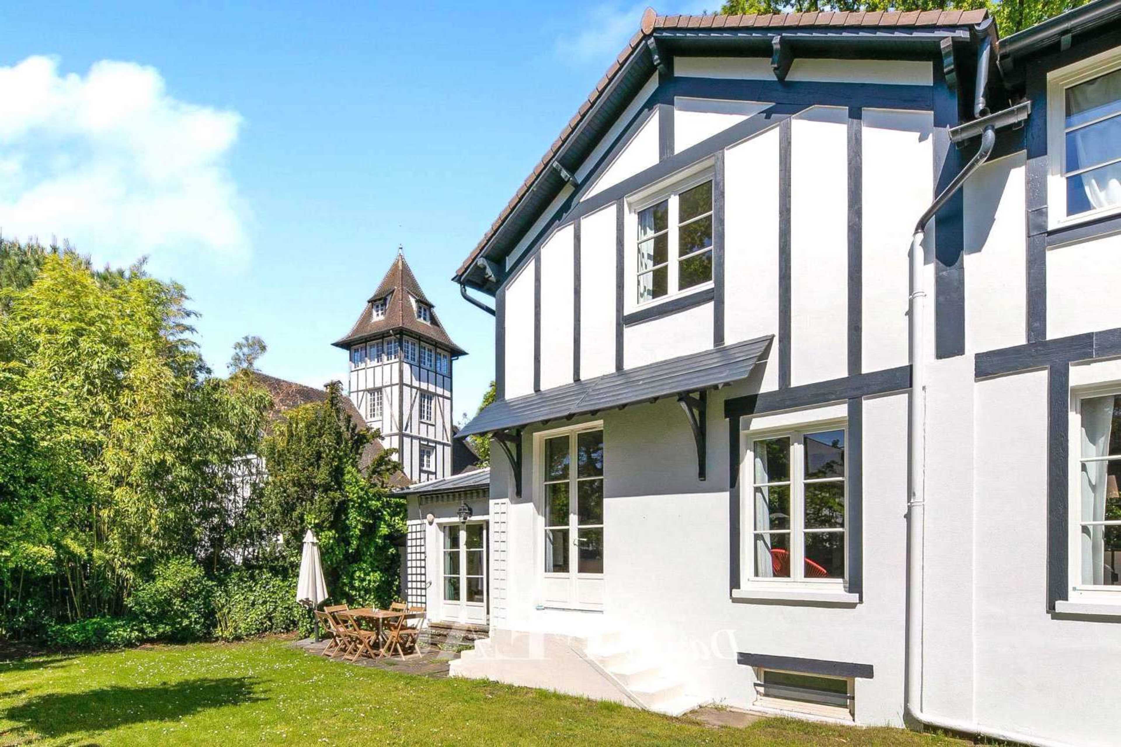 A two-story house with a white exterior and black accents, alongside a tall, pointed tower, set in a green garden with outdoor furniture.