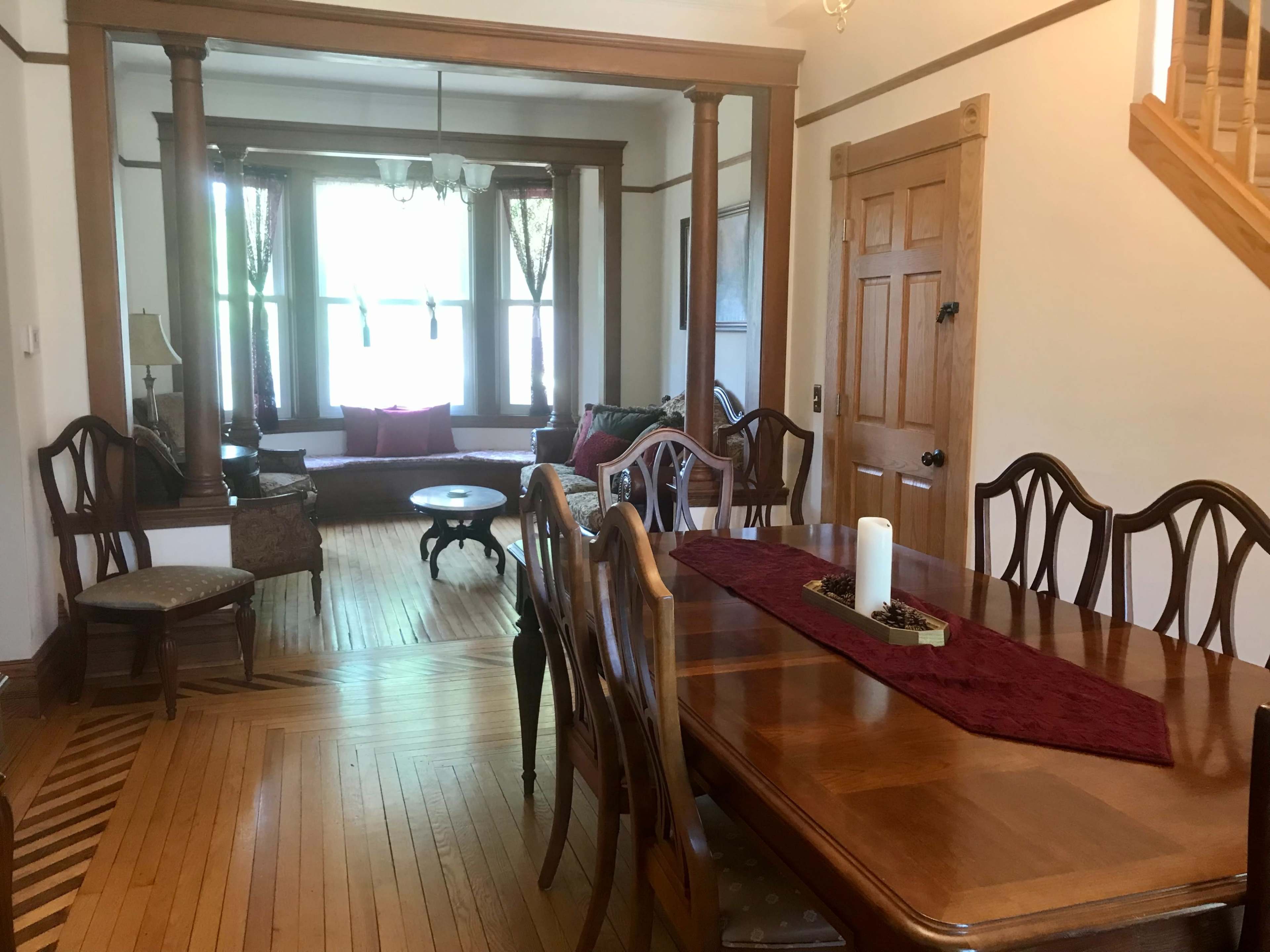 A dining area adjacent to a living room, featuring a wooden dining table set with a red runner and candle, and large windows with decorative curtains.