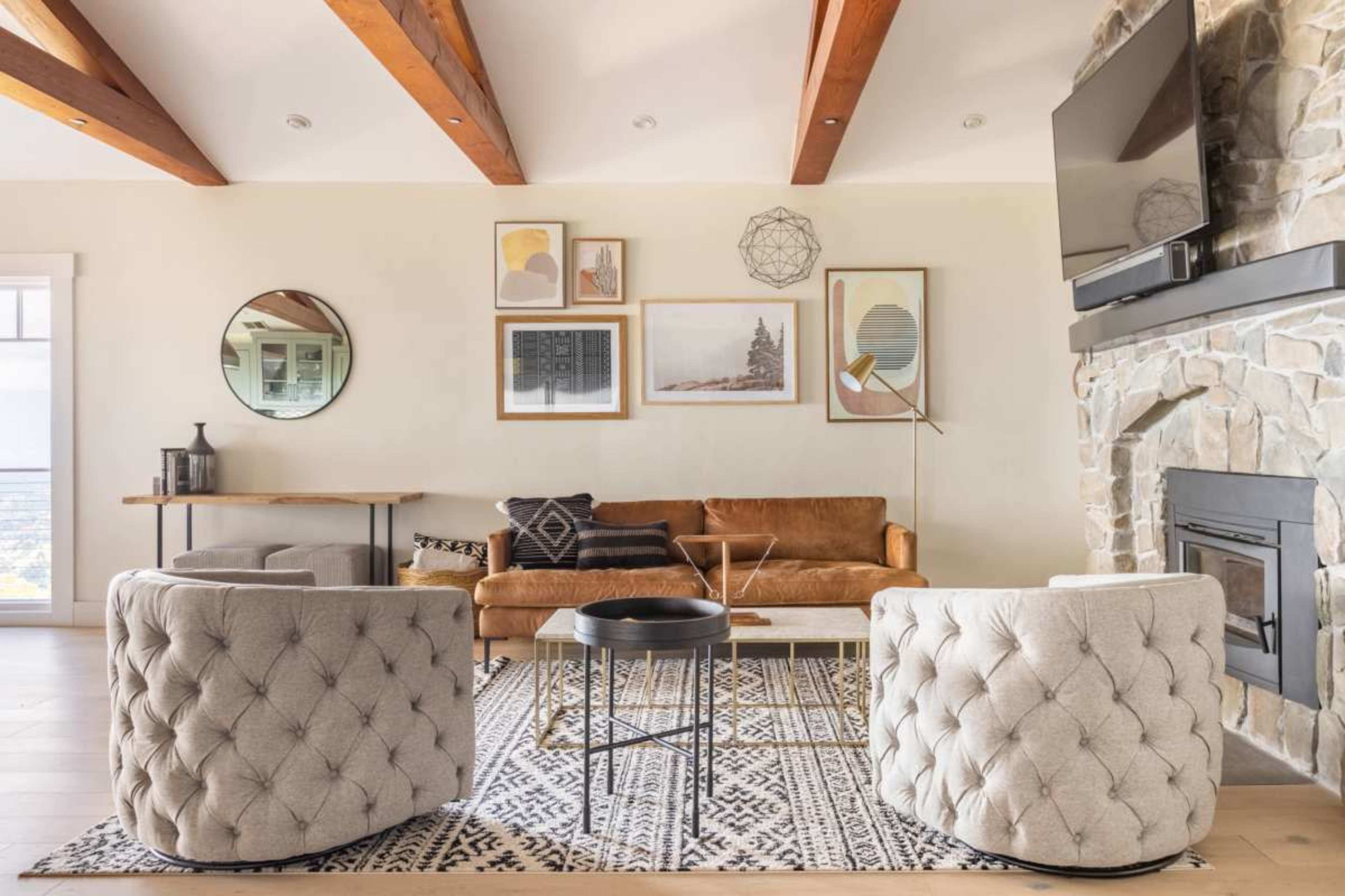 The living room features a brown leather sofa, two tufted armchairs, a round coffee table, and a stone fireplace, all arranged on a patterned rug under wooden beams.