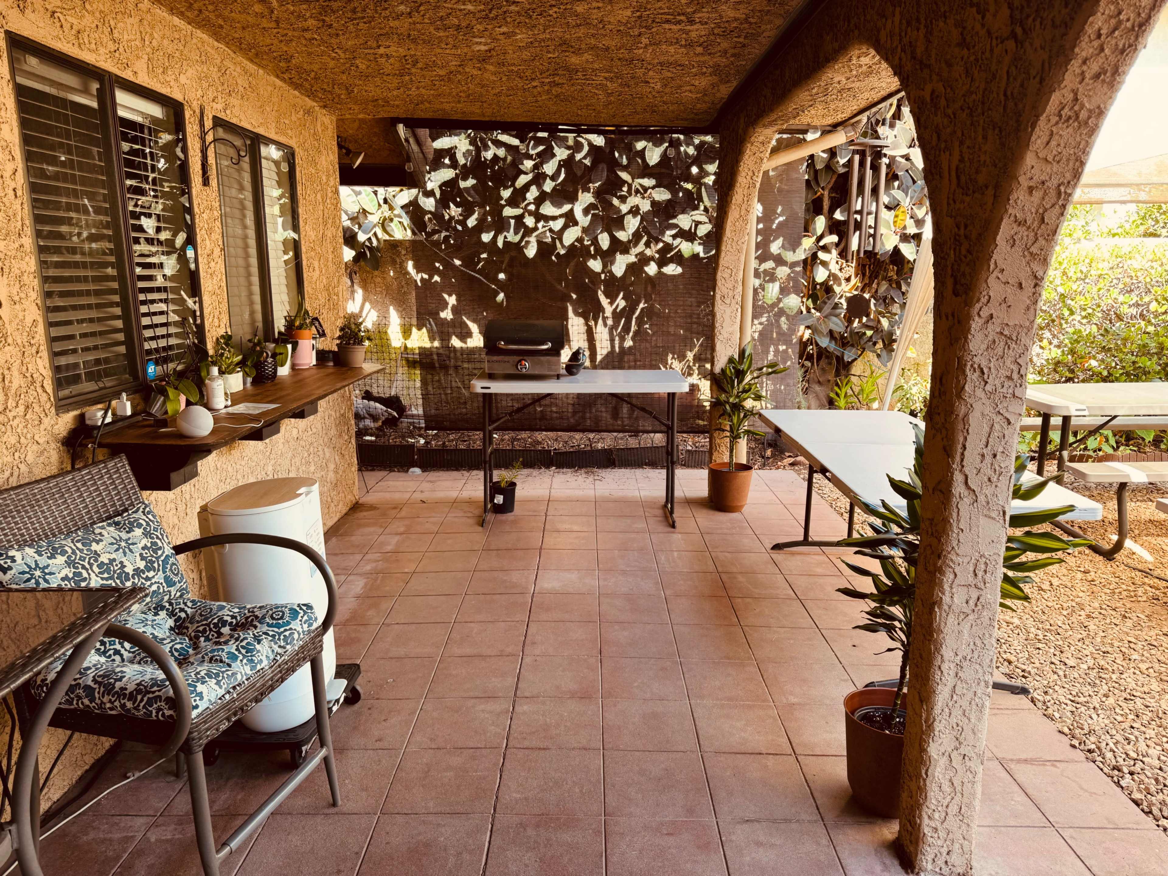 The image depicts a covered patio with tiled flooring, featuring a table with chairs, a grill, and potted plants.