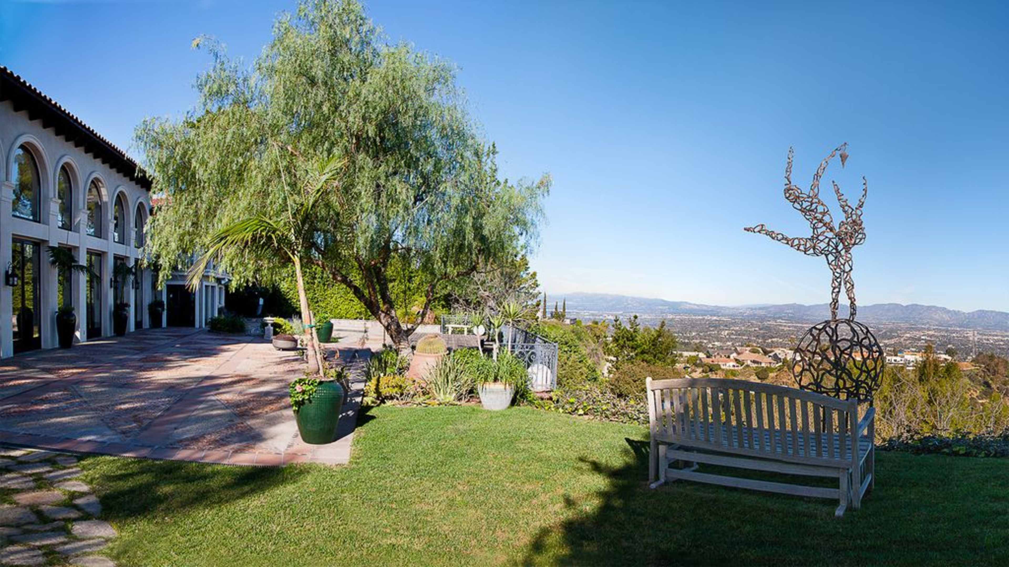 A landscaped outdoor area with a wooden bench, a sculpture, and a panoramic view of the horizon, featuring greenery and a spacious patio.