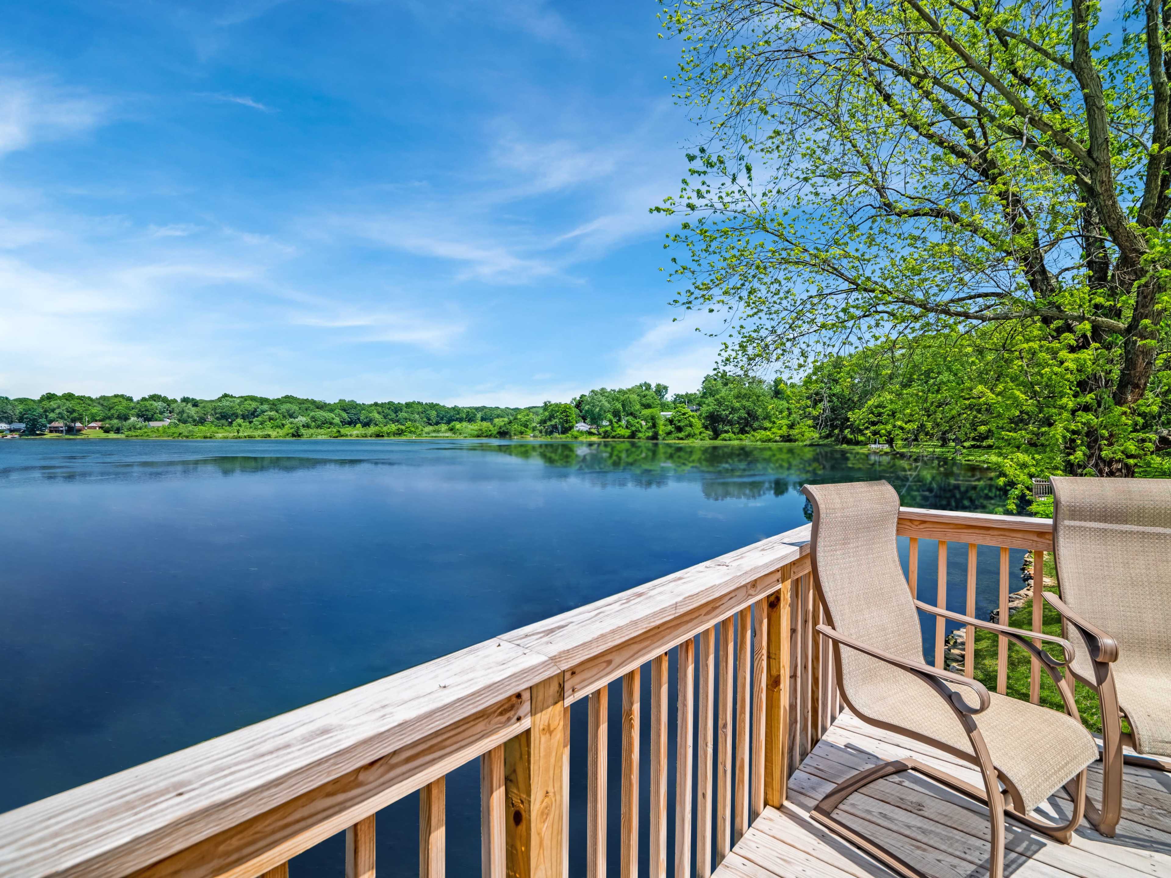 A wooden deck with two chairs overlooks a calm lake surrounded by trees.