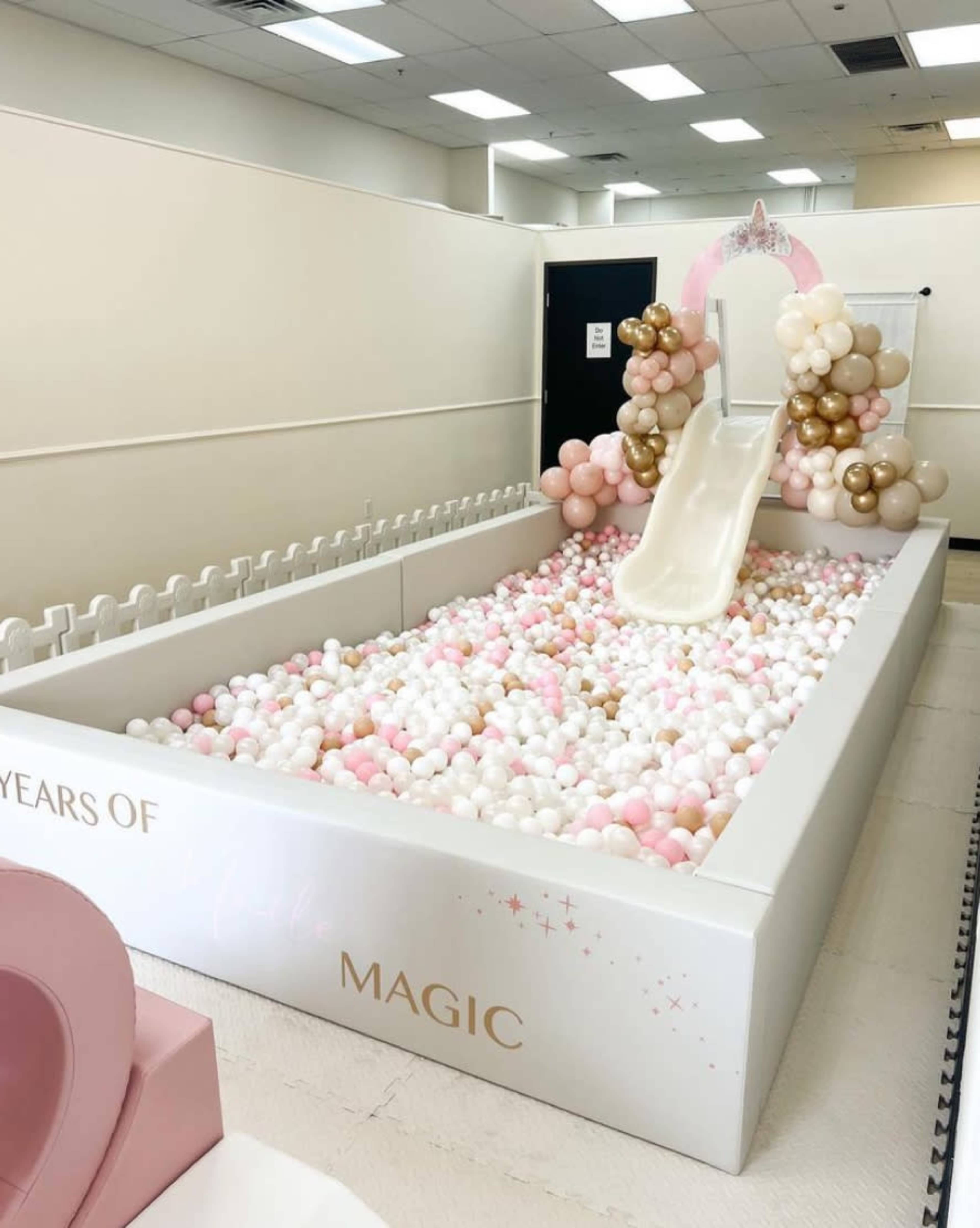 A decorated play area featuring a white slide and an abundance of colorful balloons arranged around a ball pit filled with pastel-colored balls.