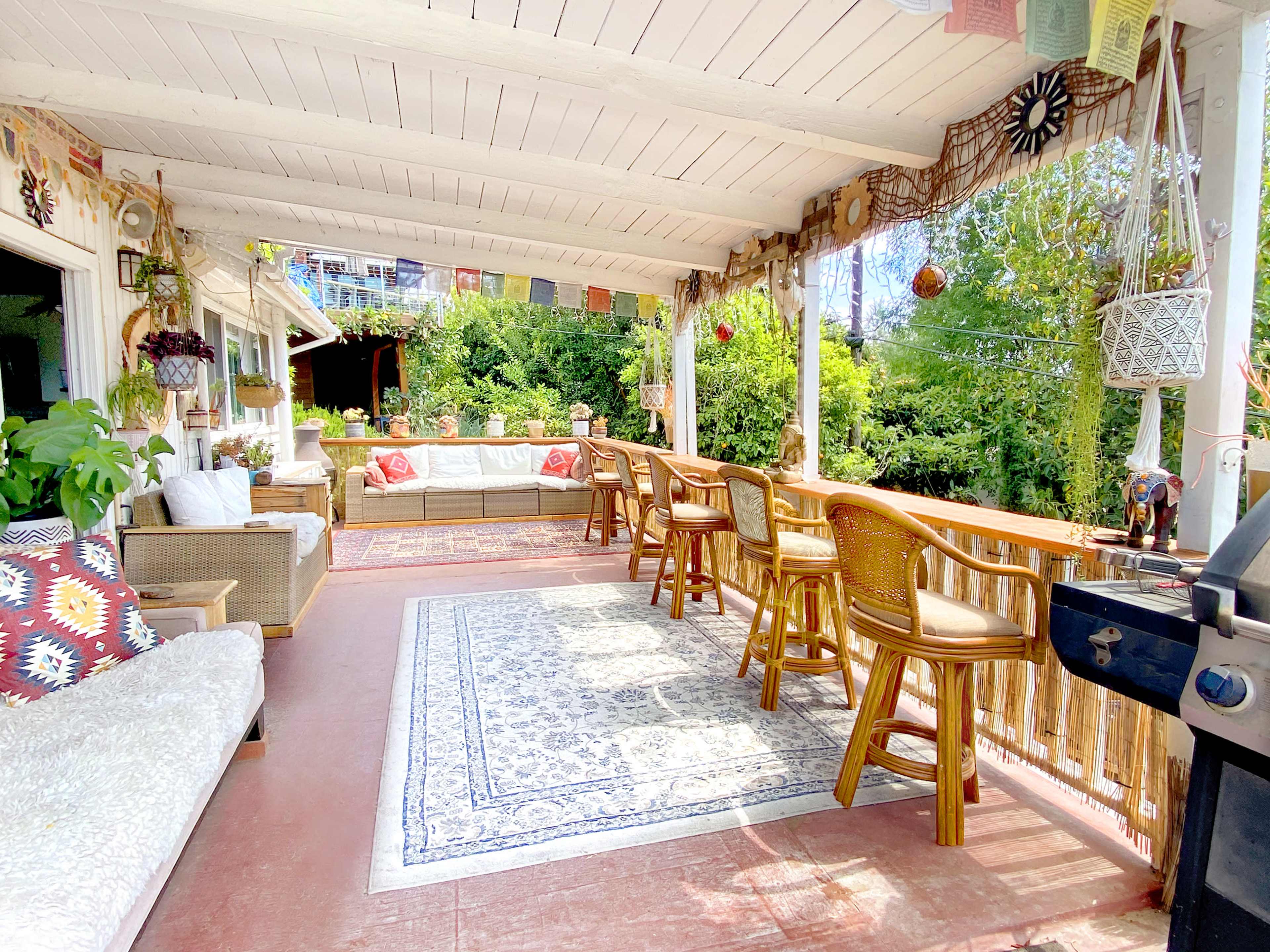 A spacious covered porch with a seating area, a bar with stools, and a barbecue grill, surrounded by greenery.
