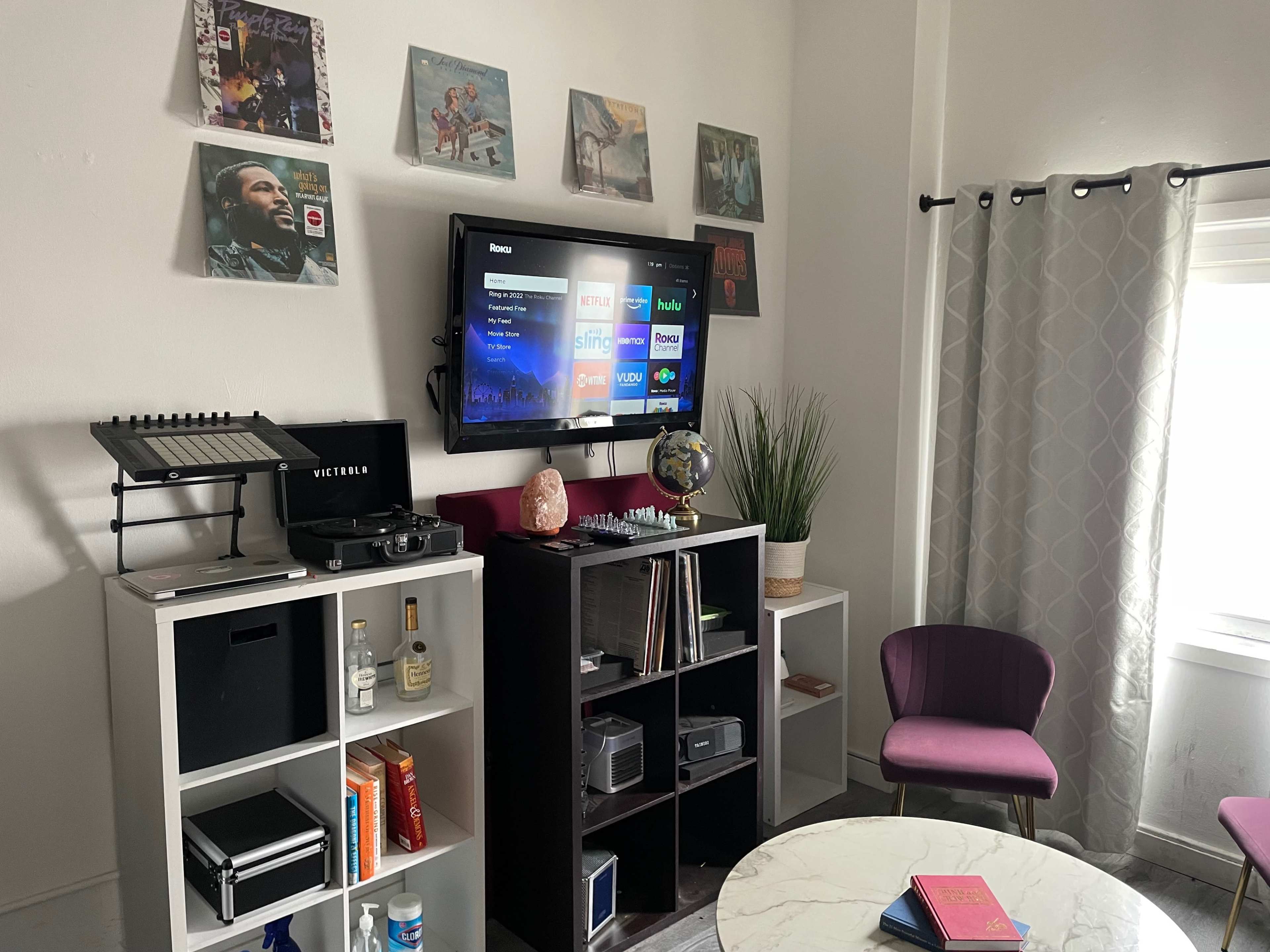 A living room corner with a television mounted on the wall above a media console, surrounded by vinyl records and decorative items, and featuring a small seating area with a round table and a purple chair.