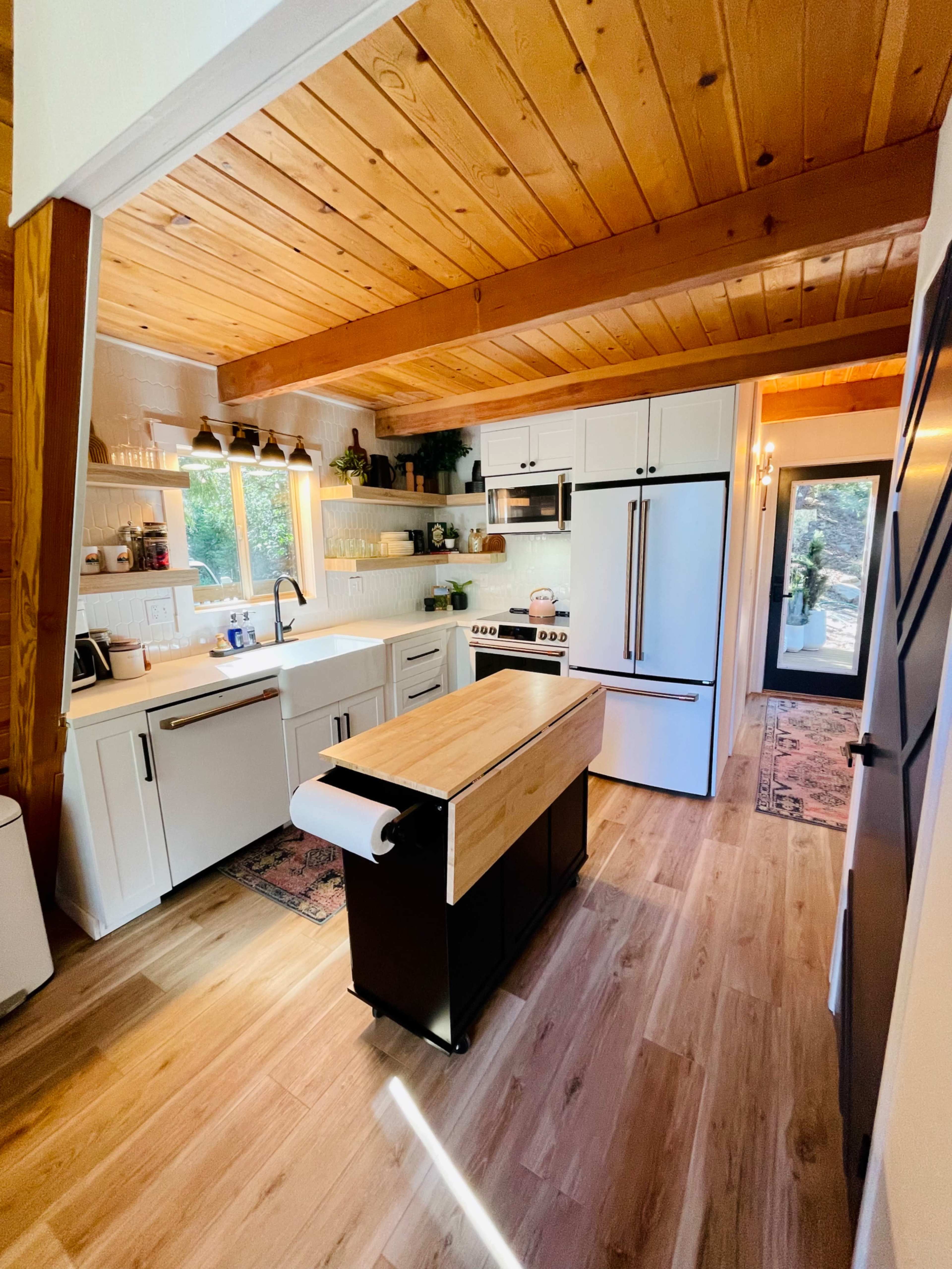 The image shows a modern kitchen with white cabinetry, a central wooden island, and large windows overlooking a natural view.