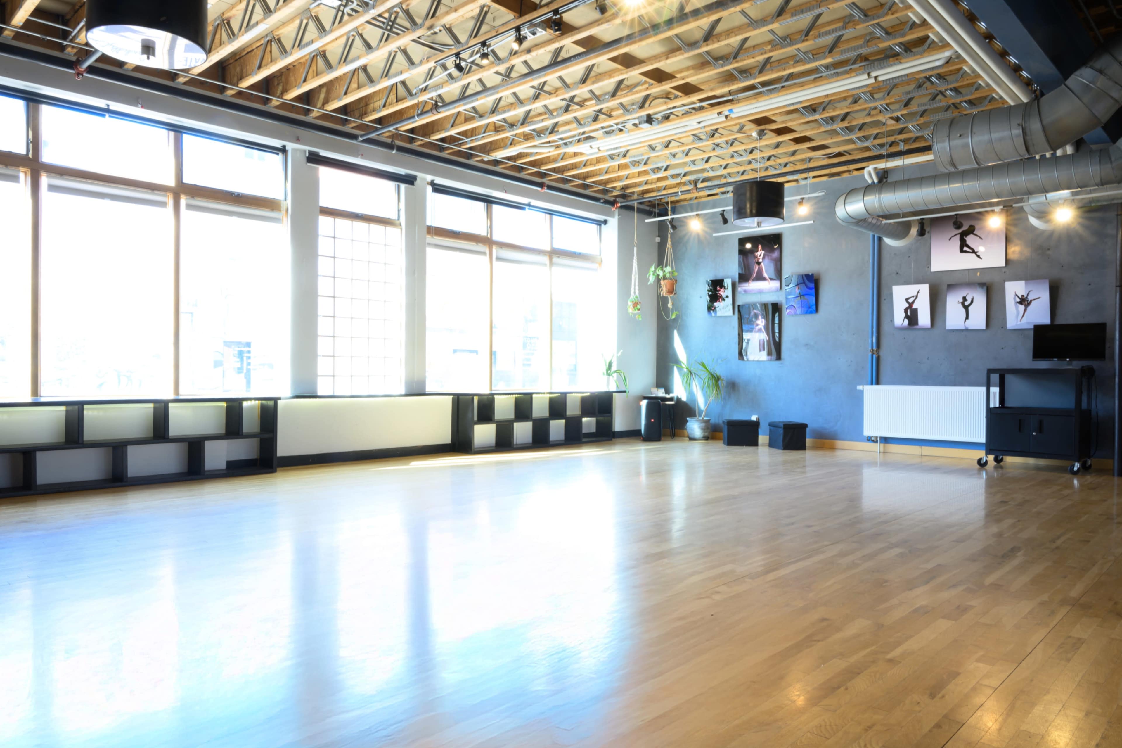 The image shows a spacious dance studio with wooden flooring, large windows allowing natural light, and walls adorned with framed dance-related art.