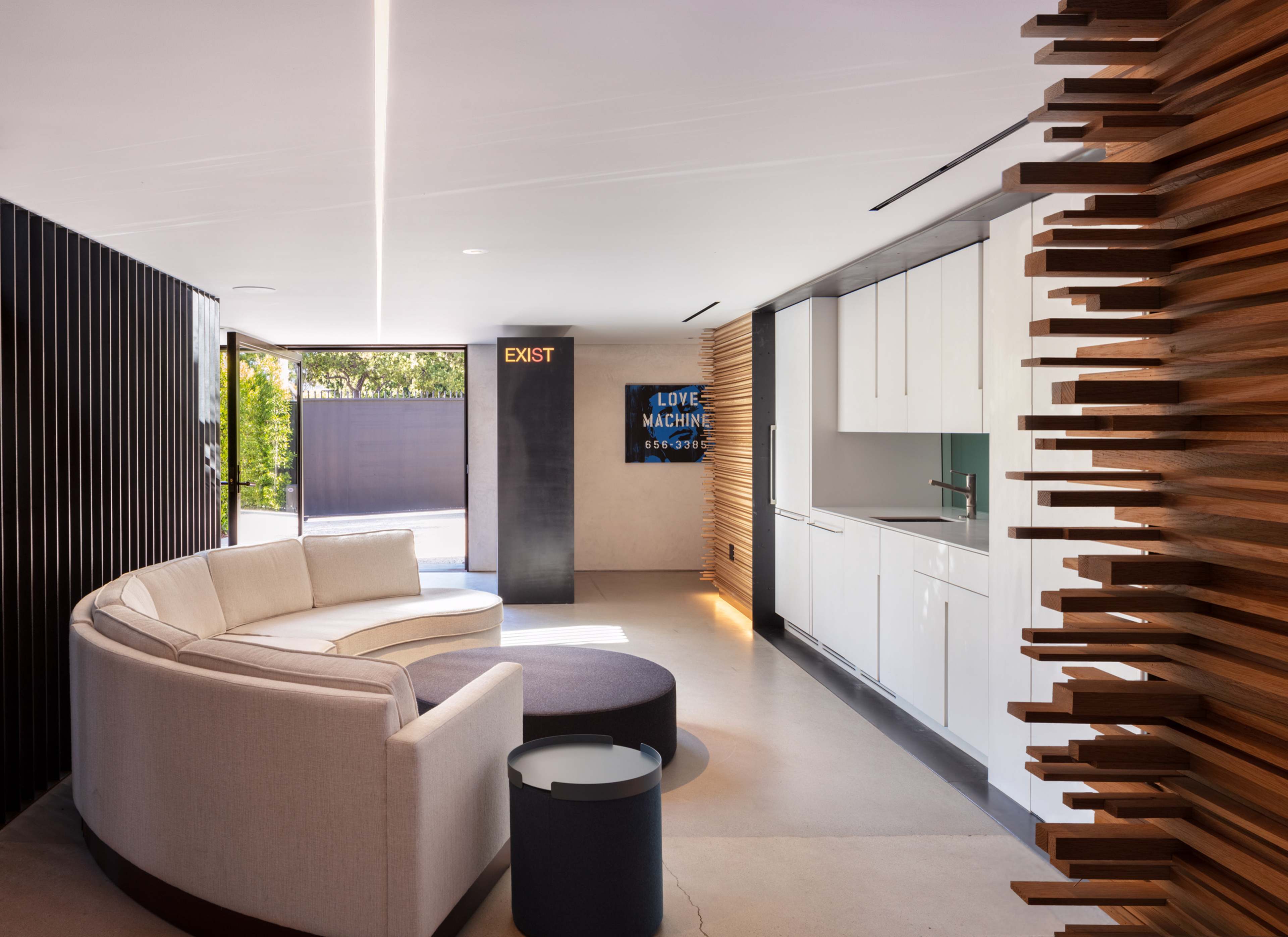 The image depicts a modern interior space featuring a curved sofa, a circular ottoman, and a kitchen area, with wooden paneling and large windows allowing natural light.