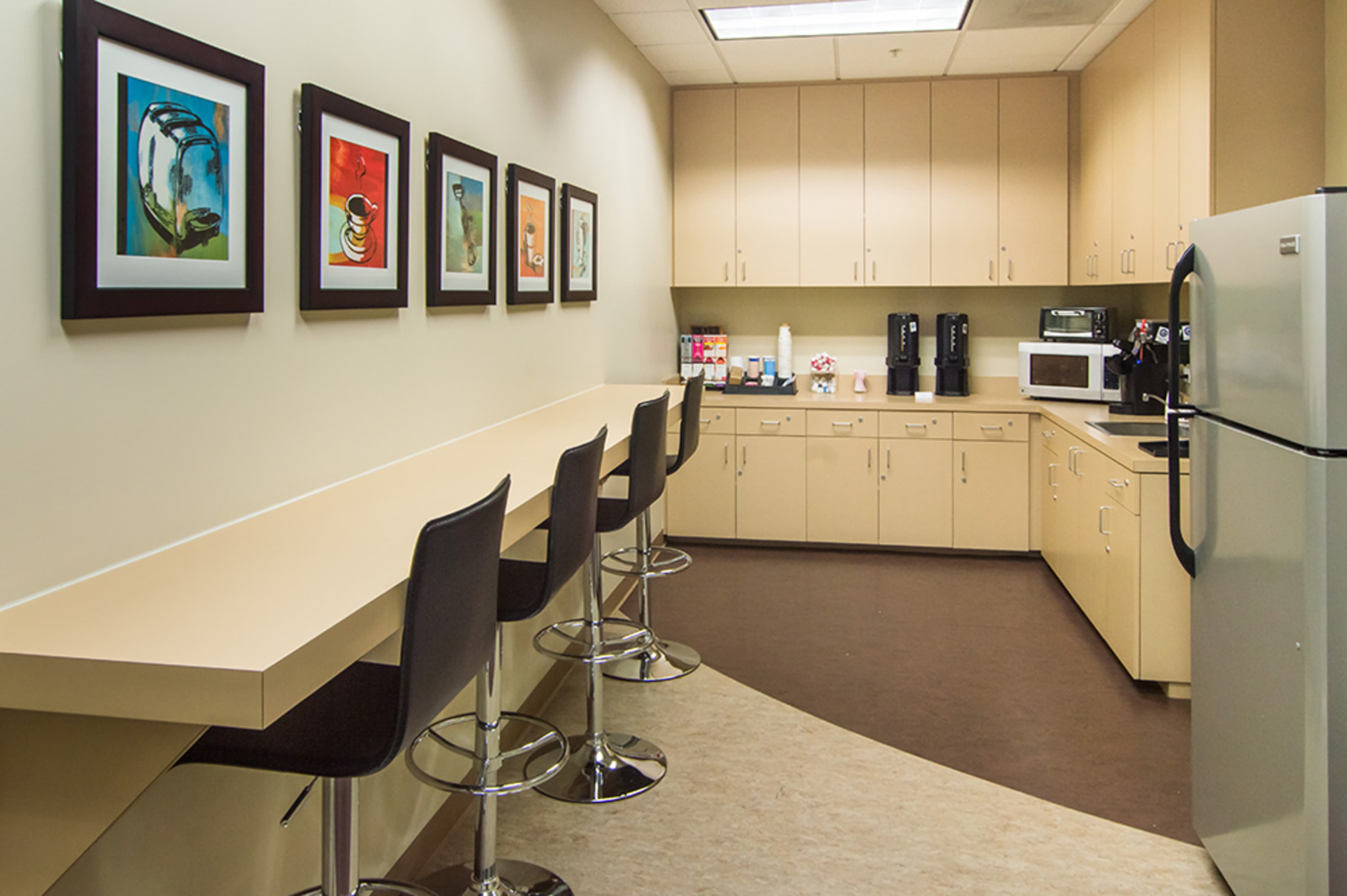 A modern break room with a long counter, bar stools, and various kitchen appliances against a cream-colored wall.
