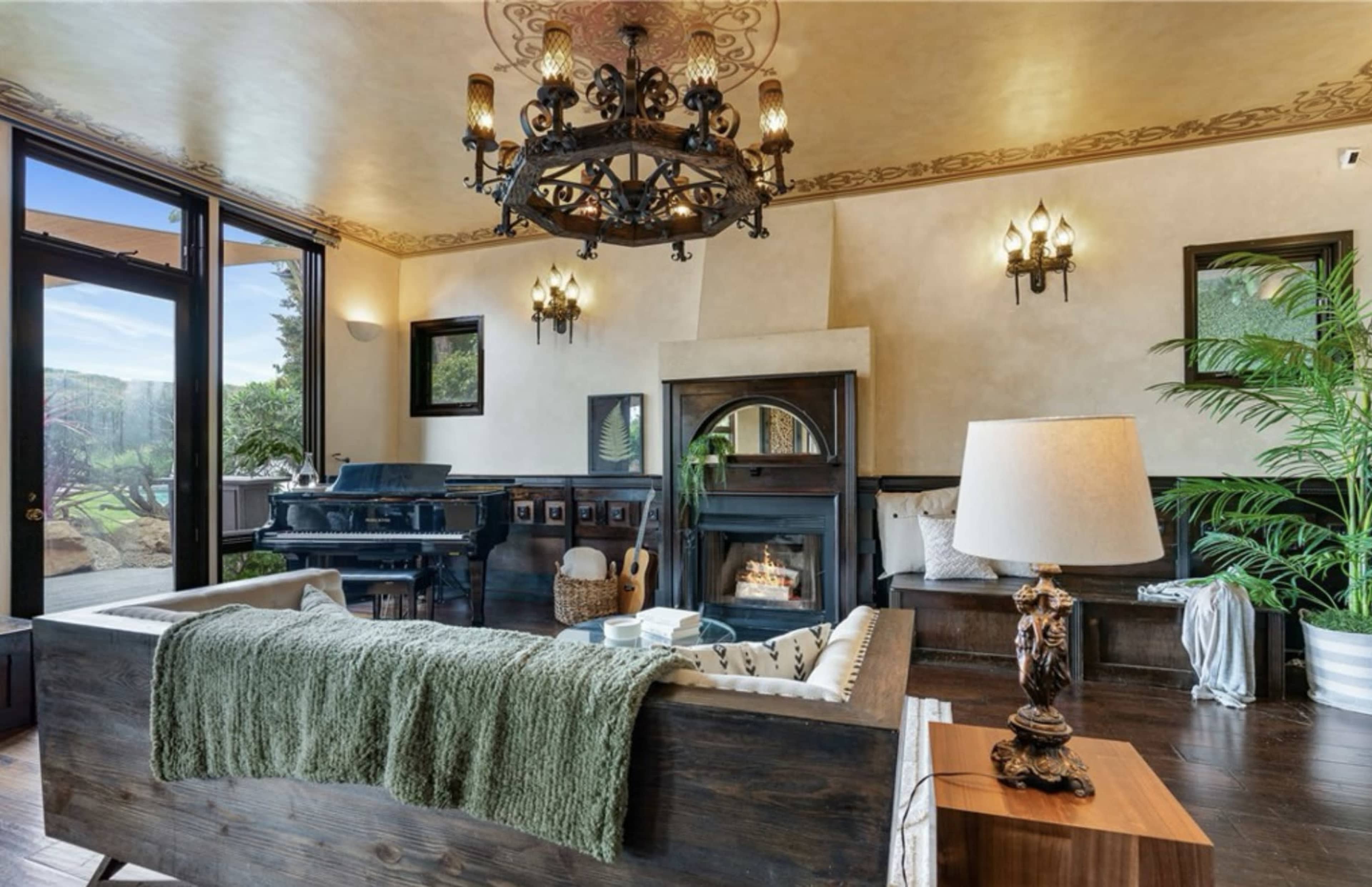 The image shows a cozy living room featuring a piano, a fireplace, and large windows that let in natural light.