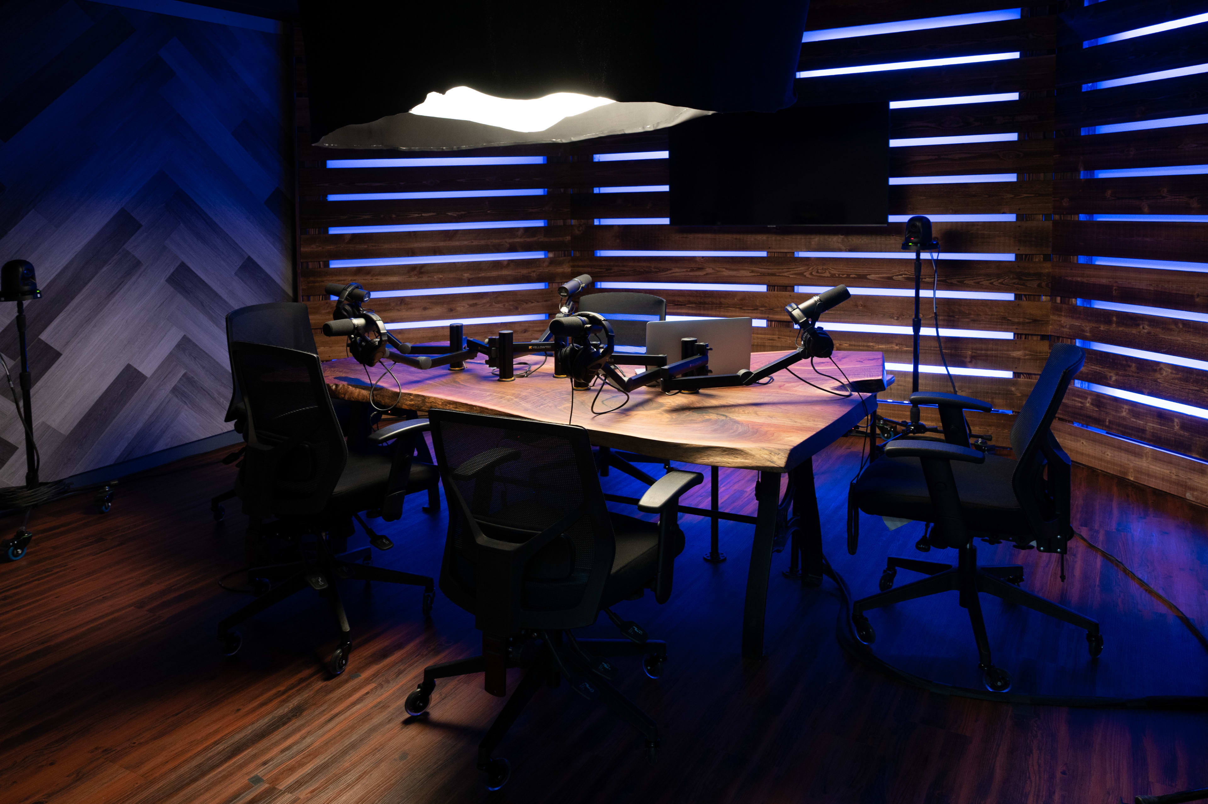 A podcast recording studio features a large wooden table surrounded by four black chairs, with microphones and equipment set up in a dimly lit space accentuated by blue lighting.