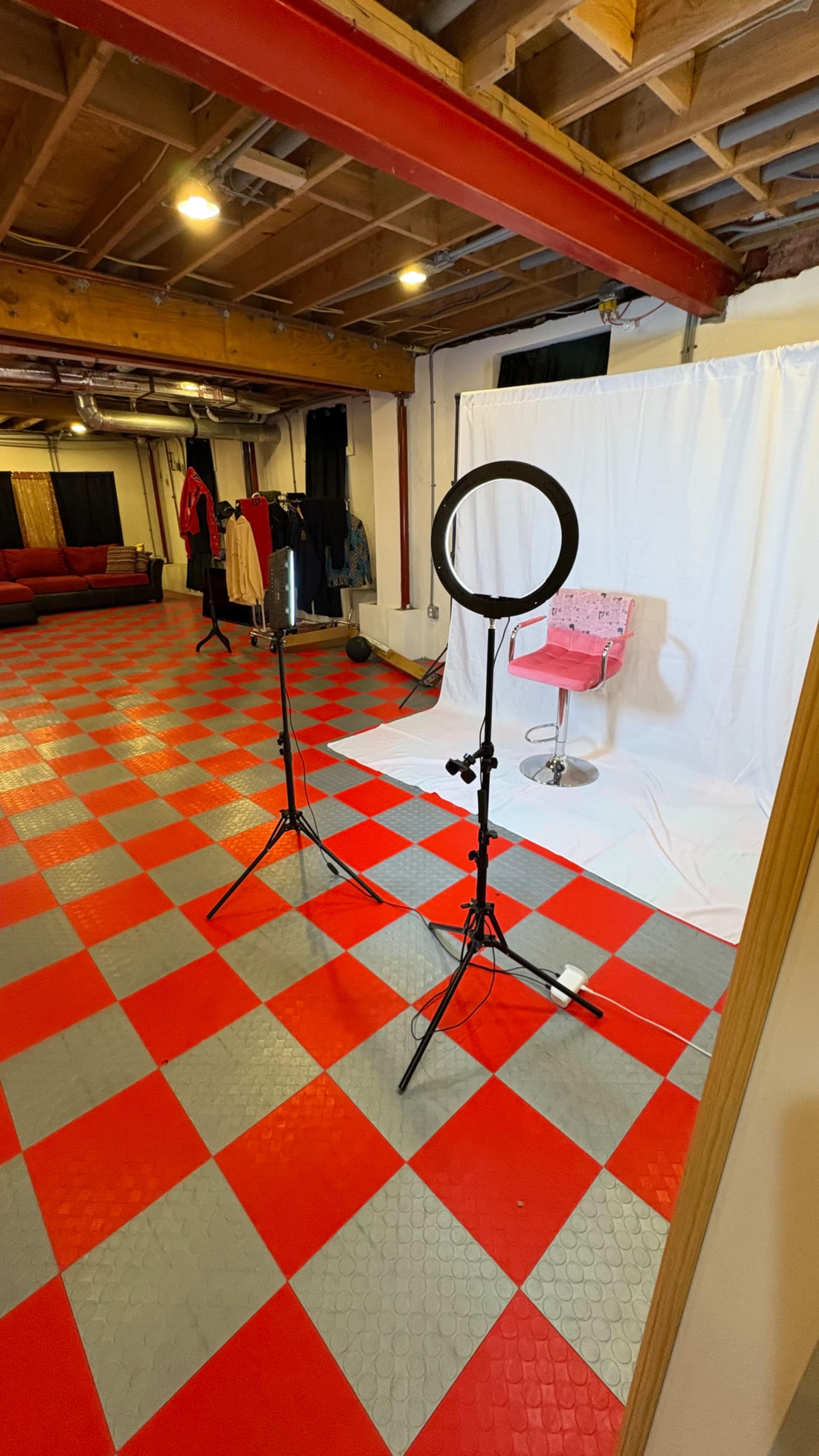 The image shows a photography studio with a red and gray checkered floor, two light stands, and a pink chair in front of a white backdrop.