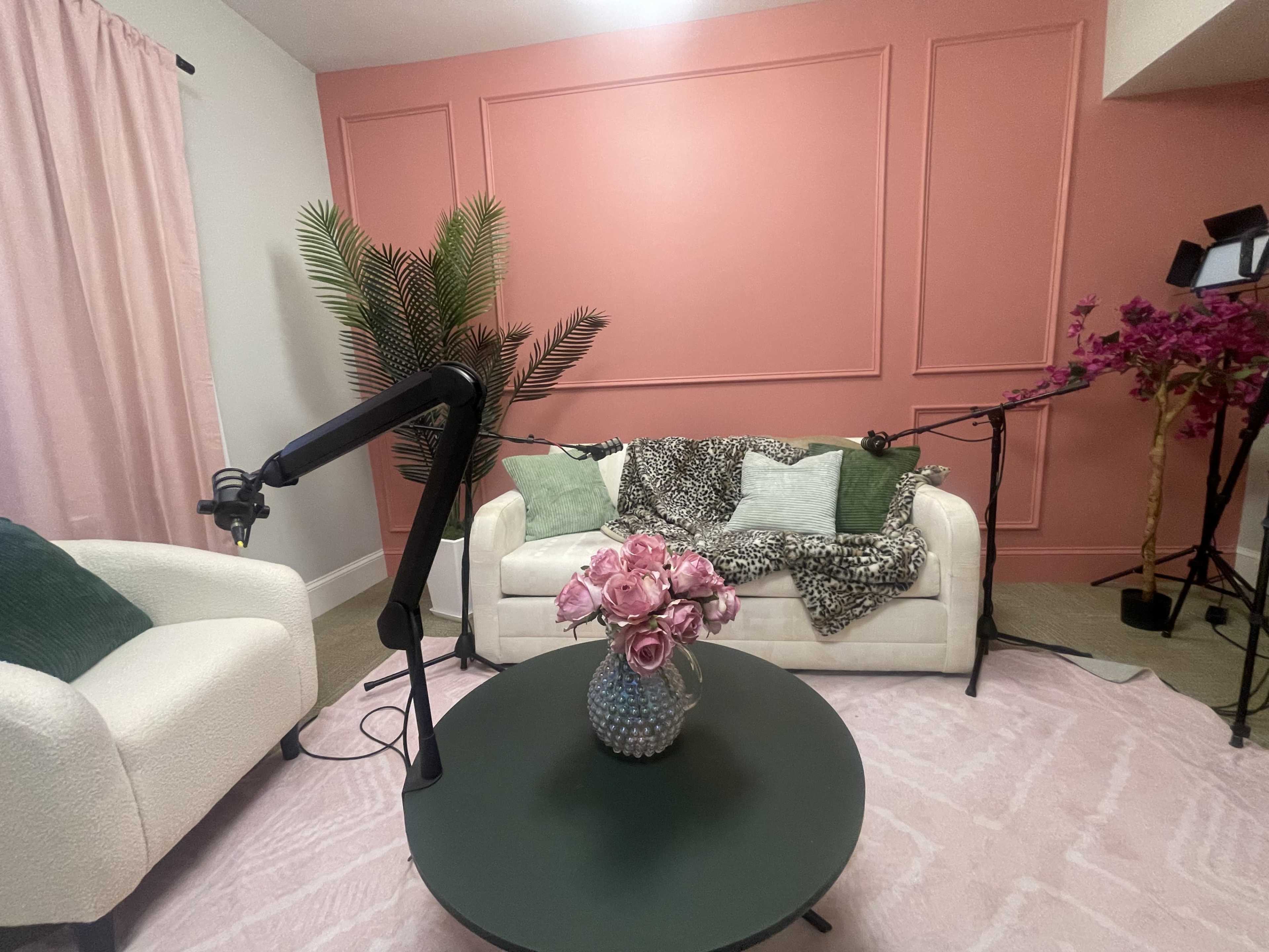 The image shows a cozy living space with a pink accent wall, a white sofa adorned with various blankets, a green coffee table, and a vase of pink flowers on top.