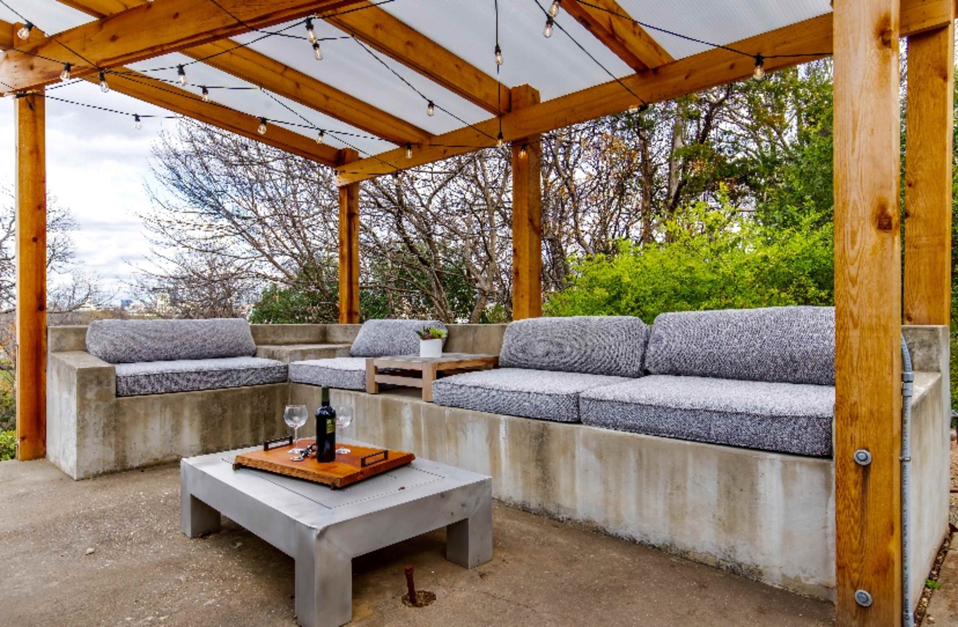 The image shows a shaded outdoor seating area with a concrete sofa, wooden beams, and a coffee table holding a wine bottle and glass.