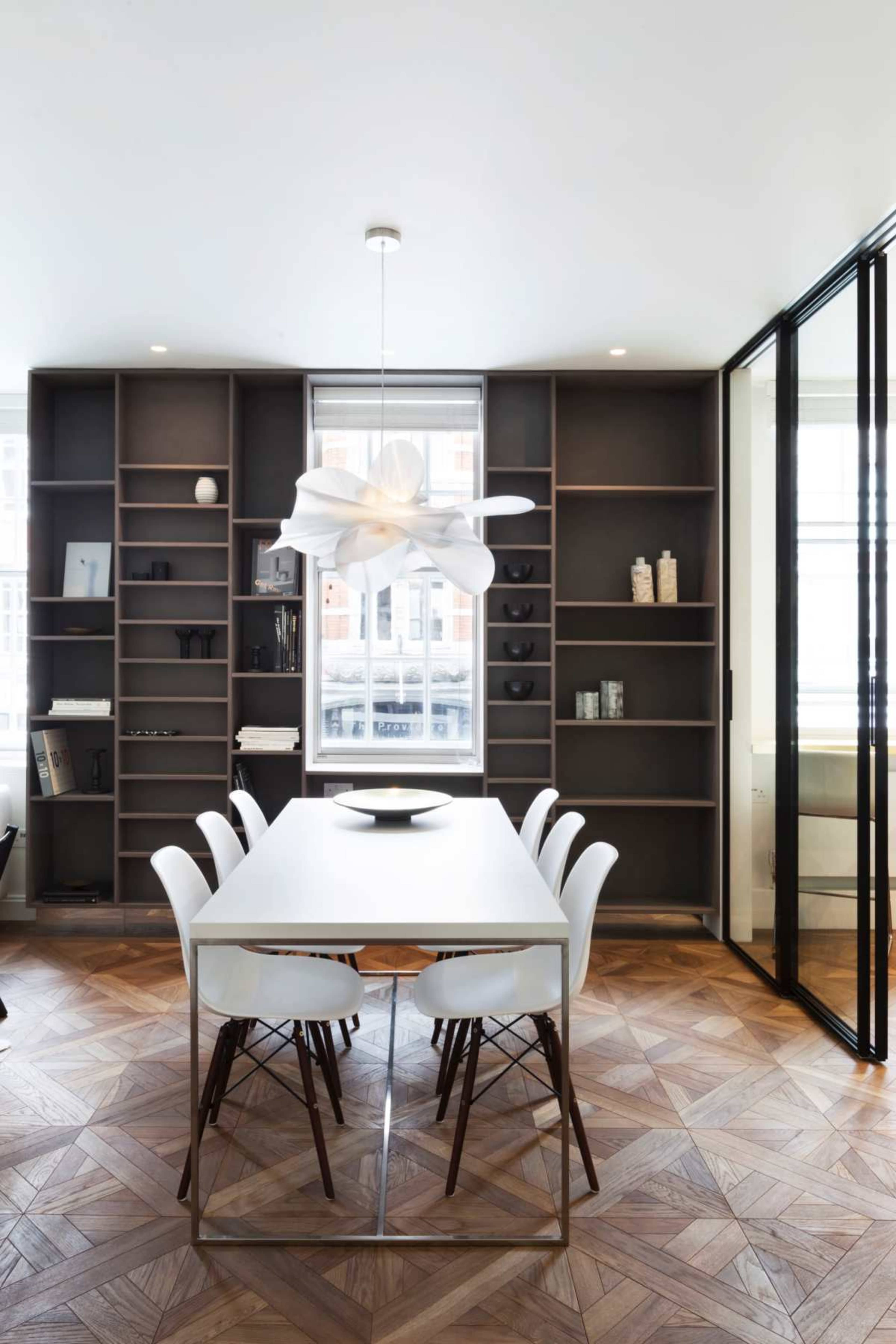 The image shows a modern dining area with a white table surrounded by six chairs, set against a backdrop of dark shelves and large windows.