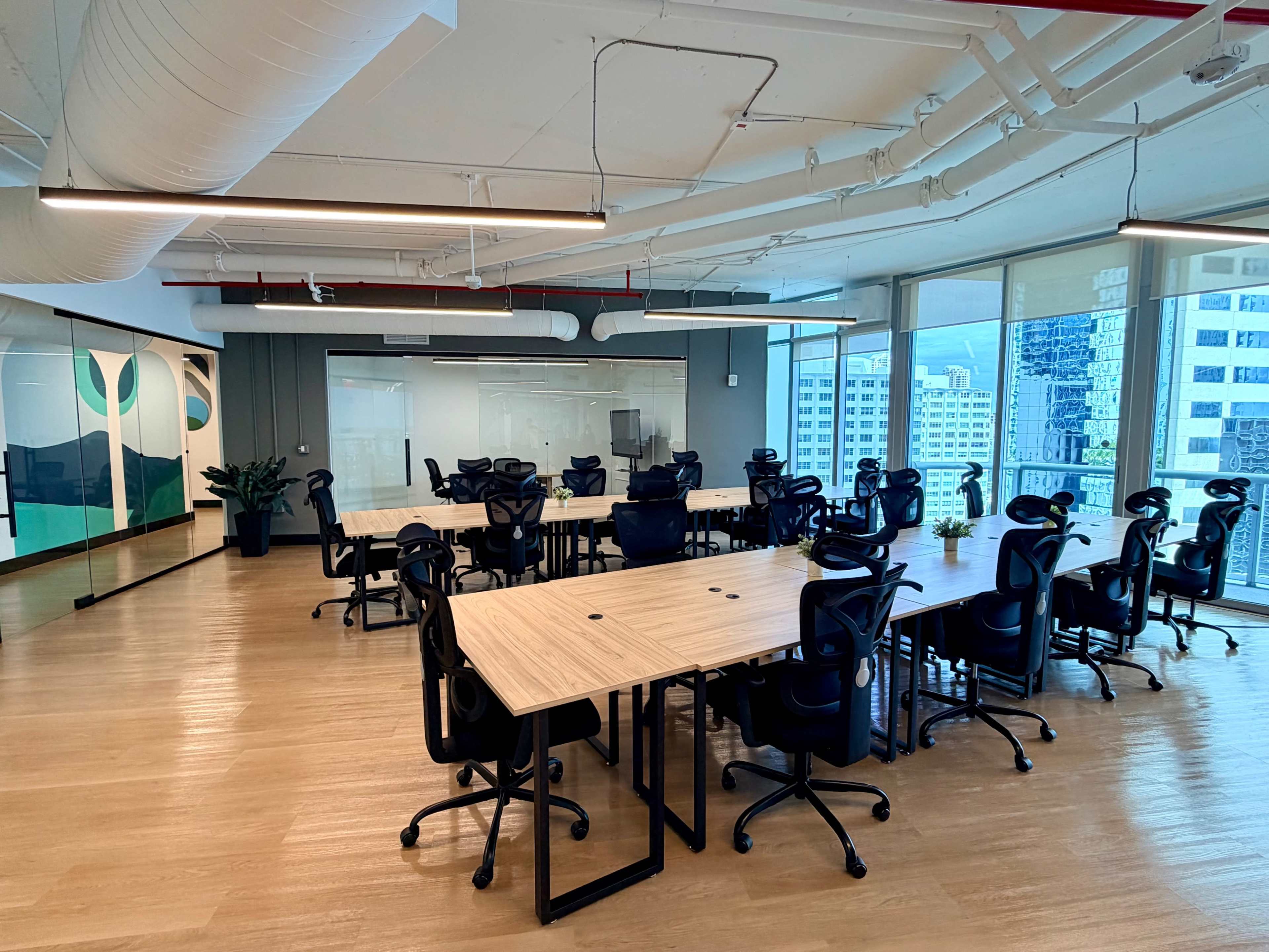 The image shows a modern office space with multiple large tables and ergonomic chairs, featuring large windows that provide a view of a cityscape.