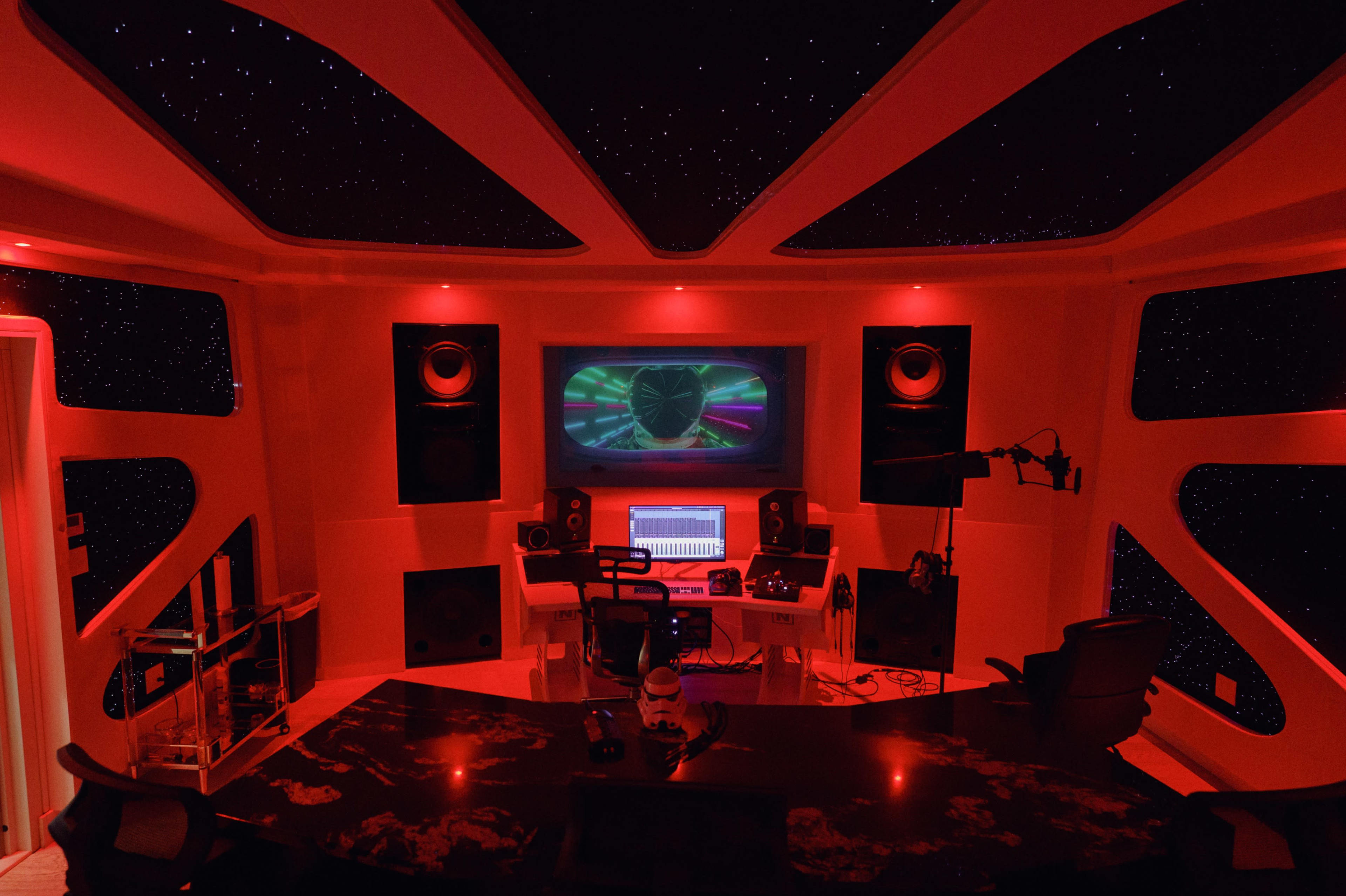 A music studio features a control desk with a computer, large speakers, and red ambient lighting, all set against a starry ceiling backdrop.