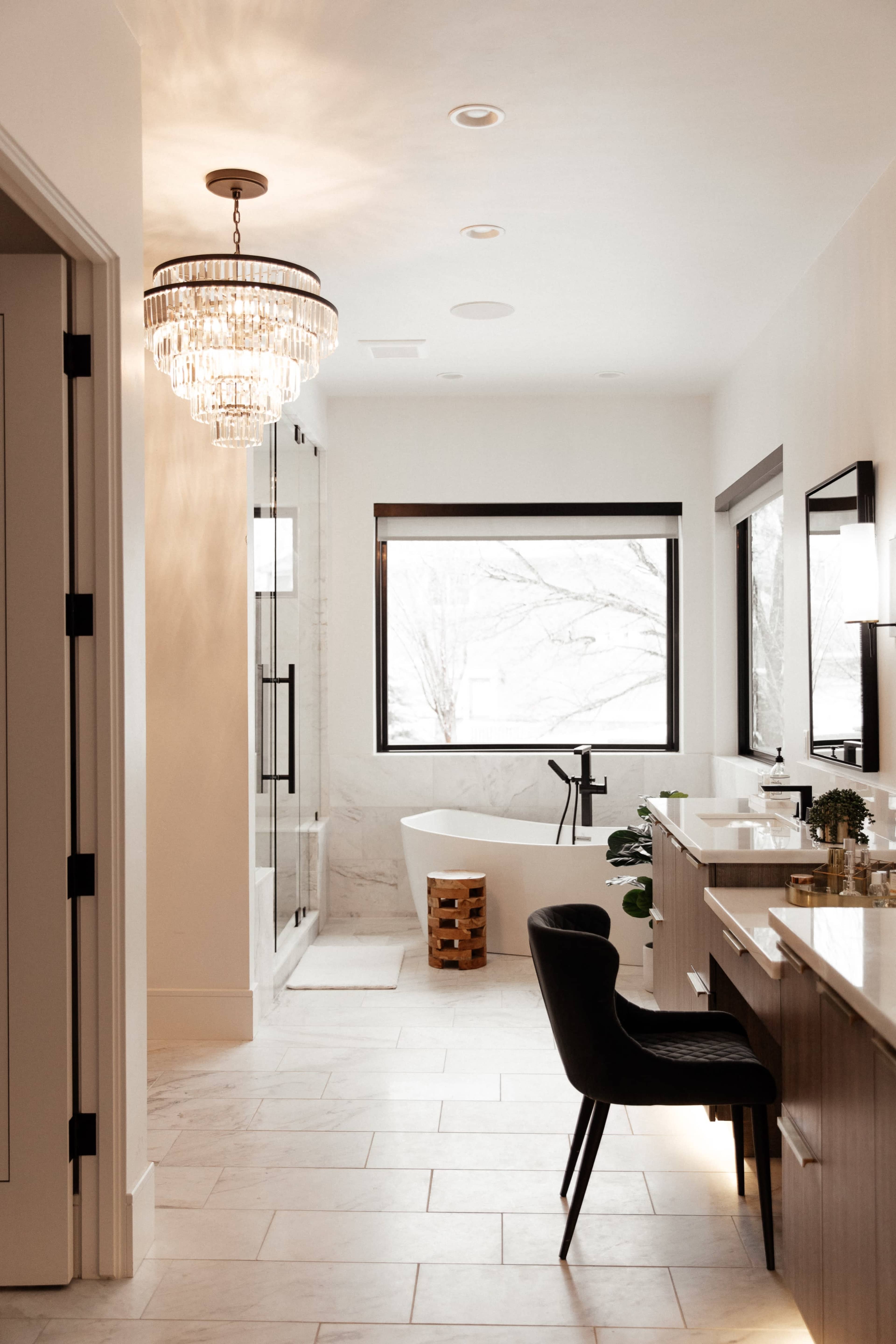 A modern bathroom features a freestanding tub, a glass-enclosed shower, and a chandelier, with ample natural light coming through large windows.