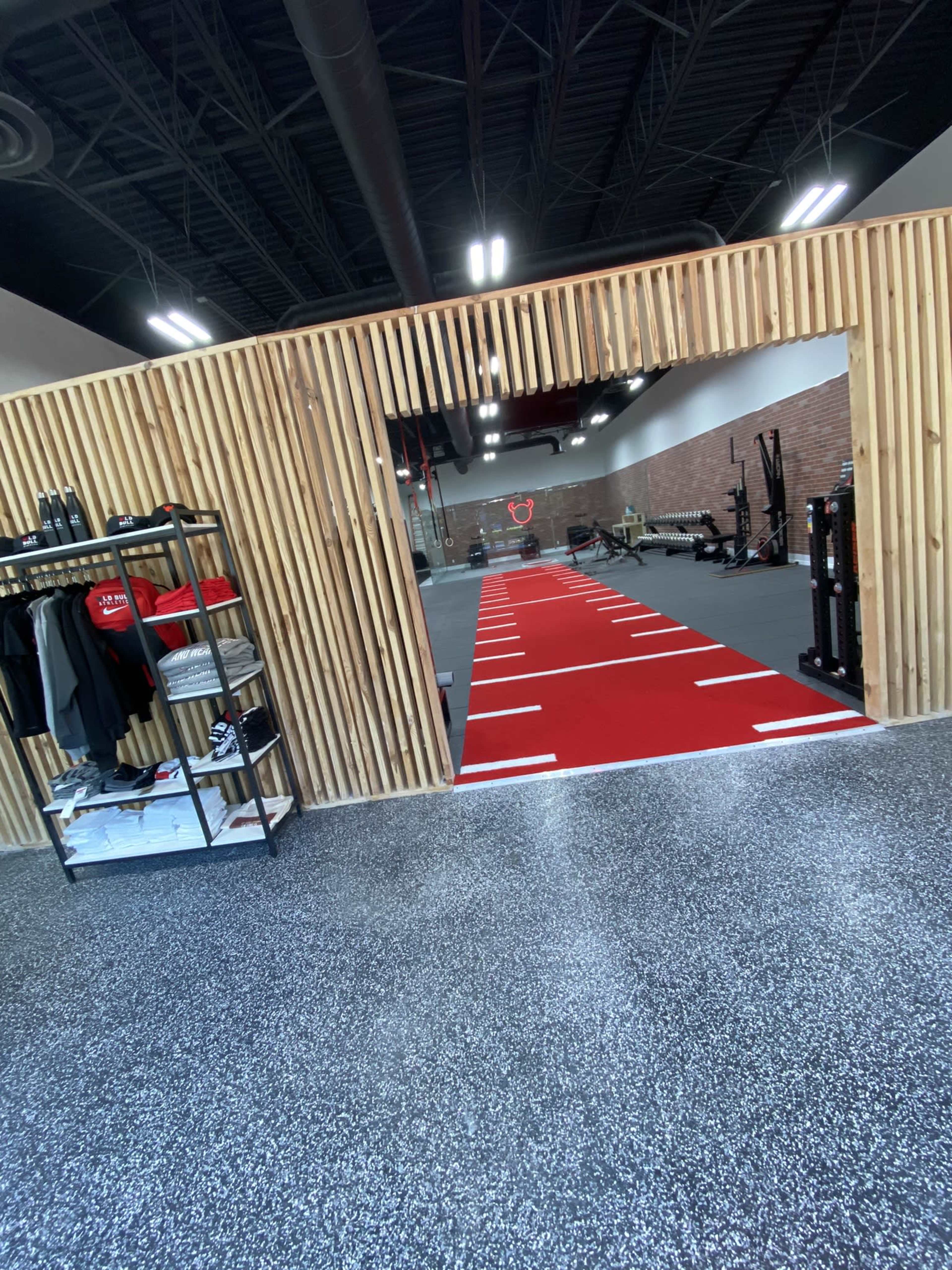 A modern gym interior with a red track leading through a wooden archway towards various fitness equipment in the background.