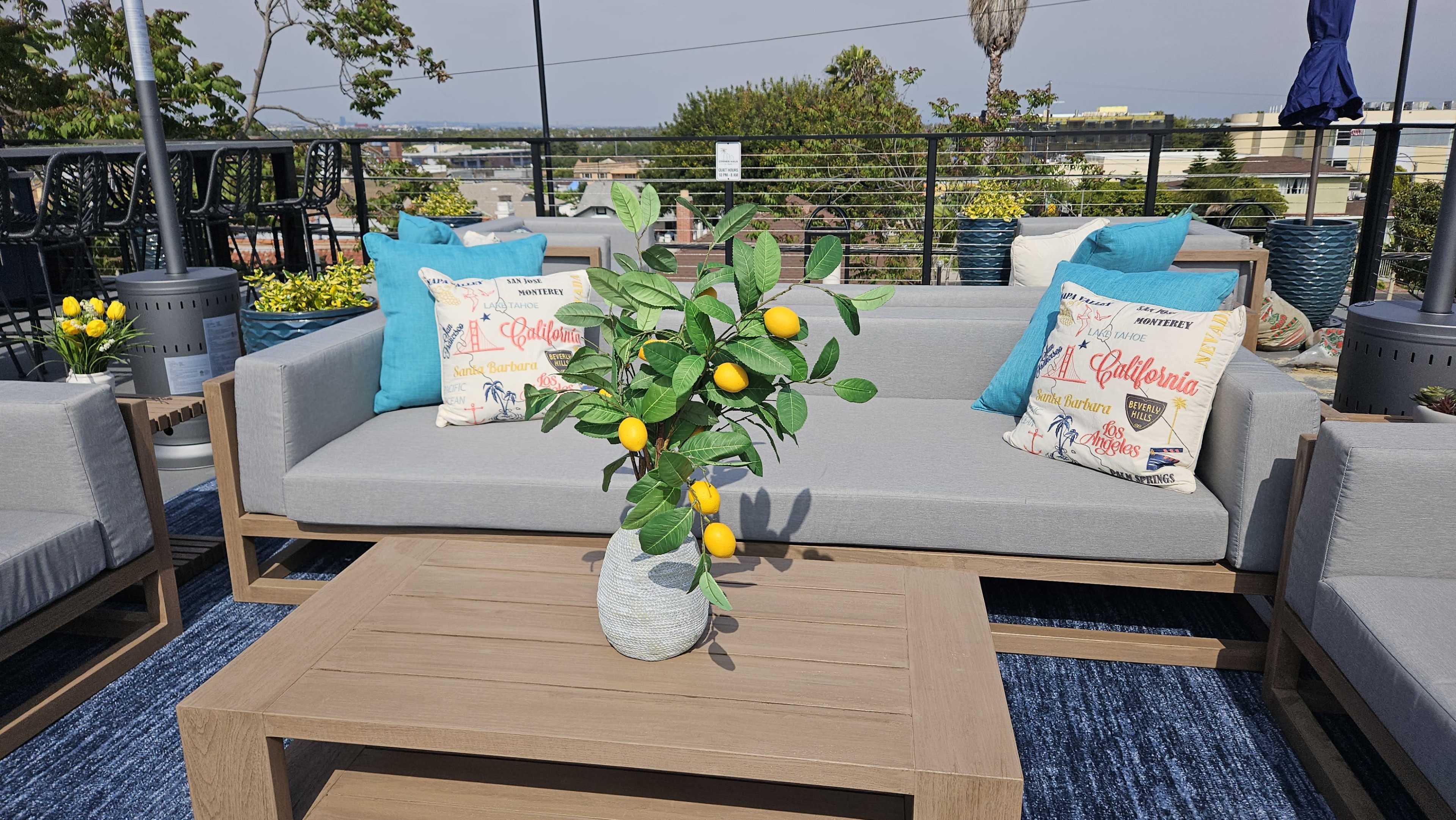 A modern outdoor seating area features gray sofas adorned with decorative pillows and a table holding a vase with a lemon plant, set against a backdrop of a cityscape.