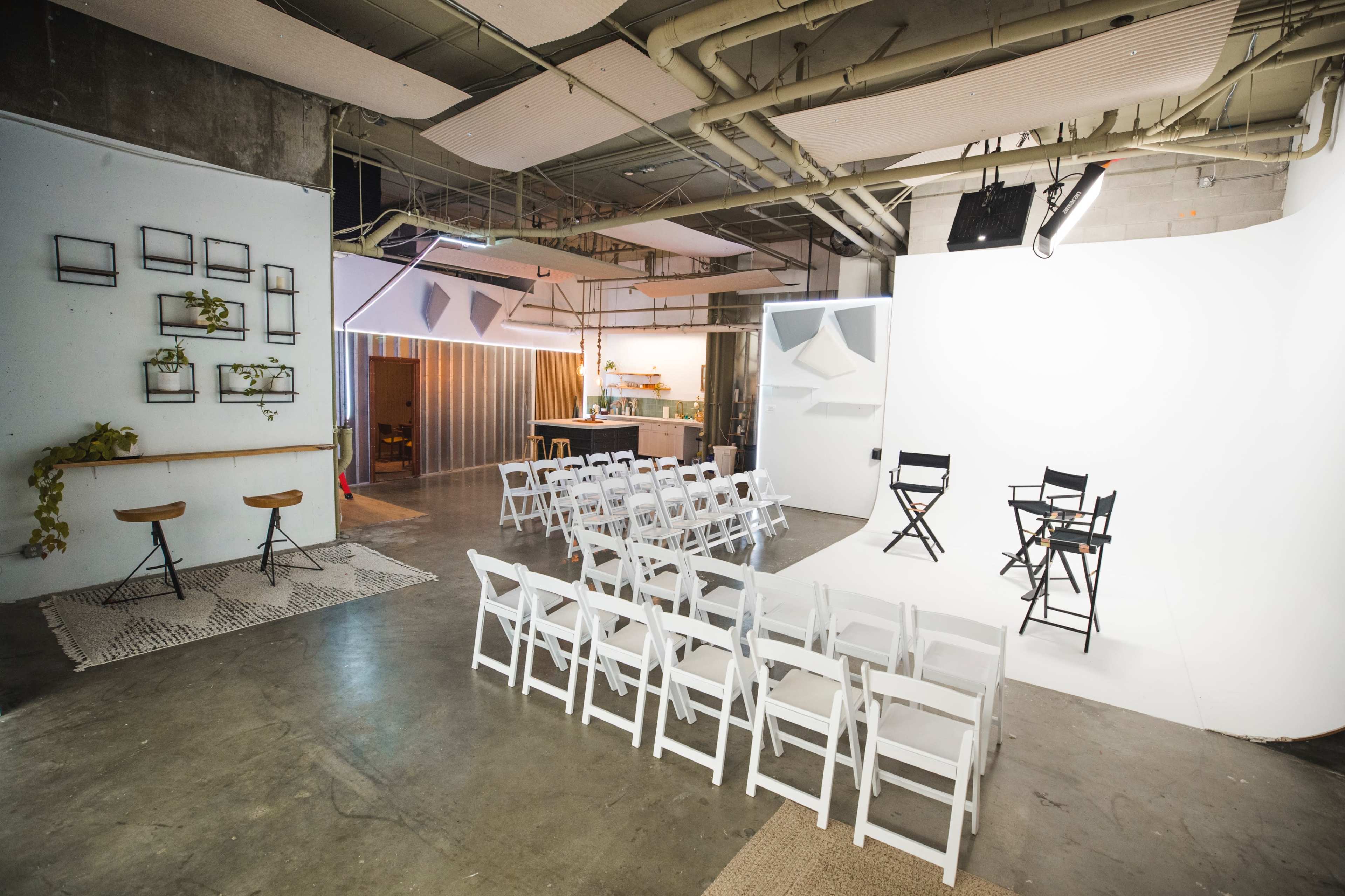 The image shows a minimalist studio setup with white chairs arranged in rows facing a blank backdrop, accompanied by two director's chairs positioned on a white platform.