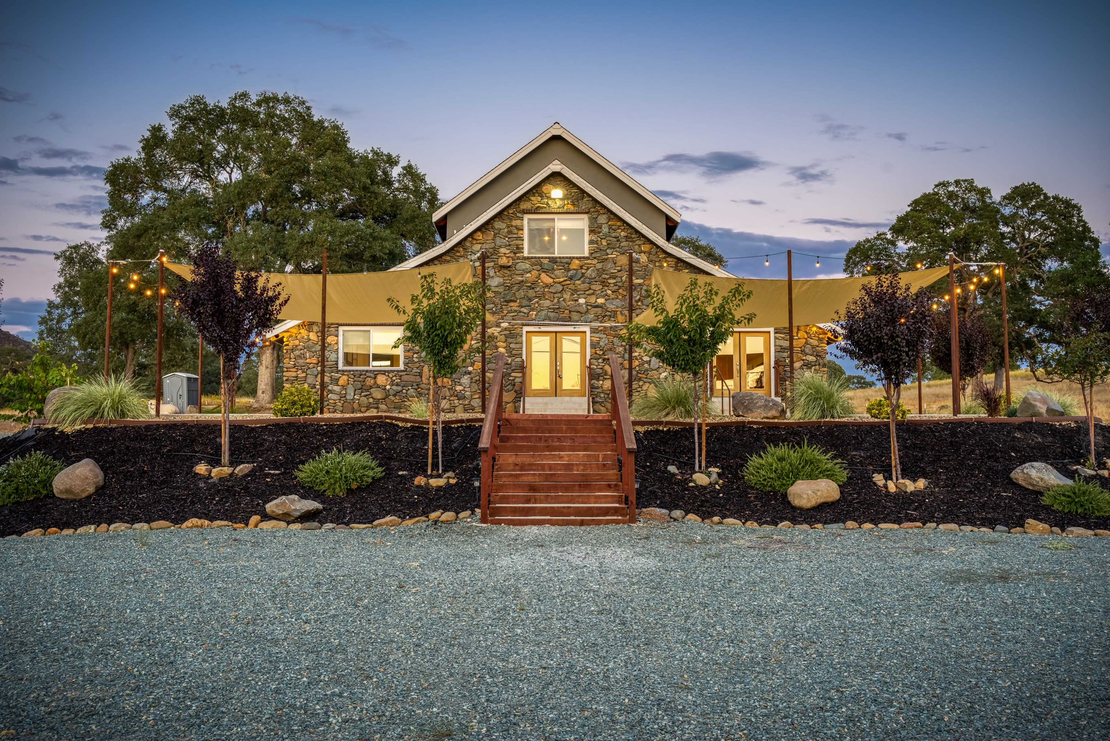 A stone house with a staircase and outdoor canopy is set against a twilight sky, surrounded by landscaped plants and gravel pathways.