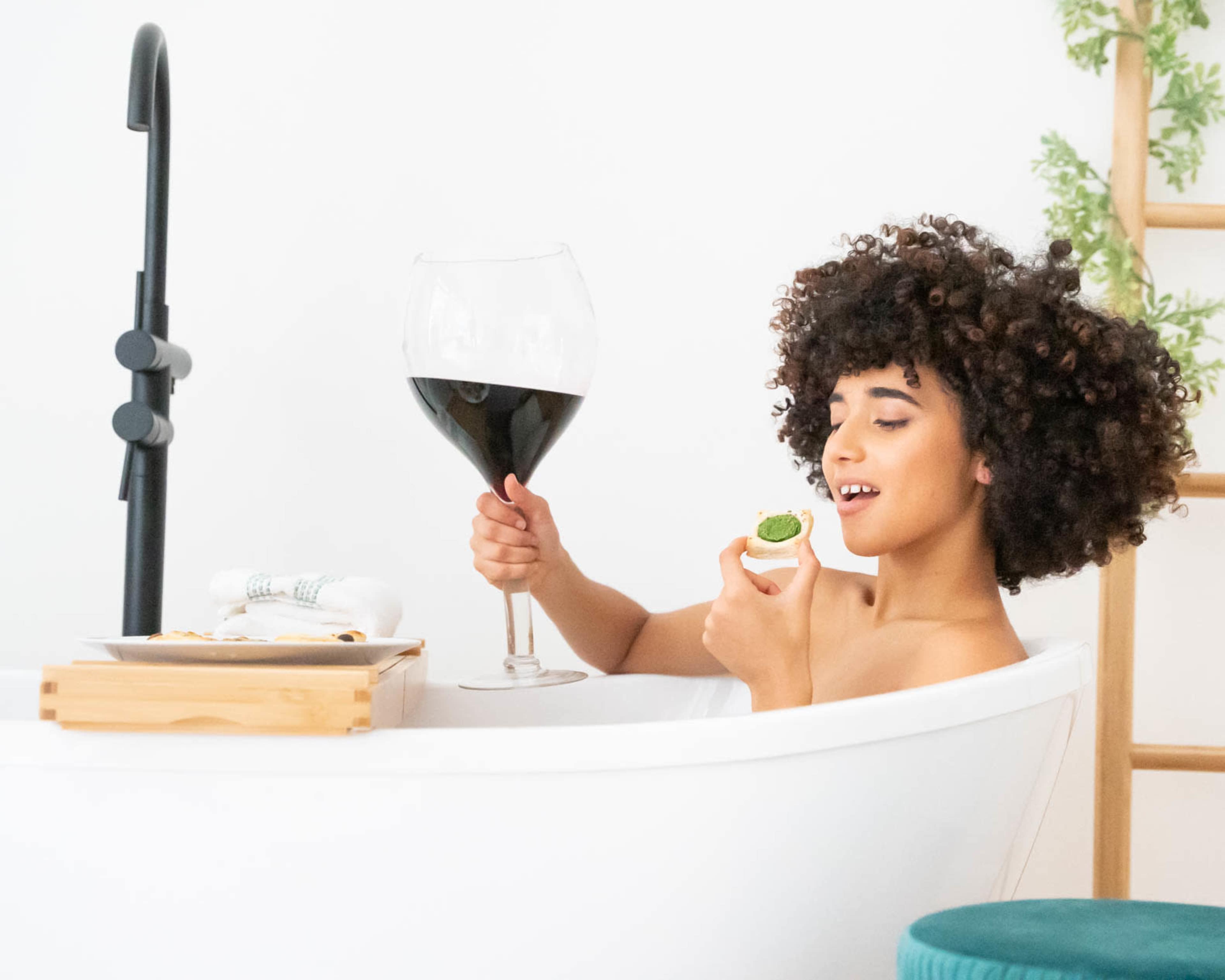 A person with curly hair sits in a bathtub holding a large glass of red wine and a small bite-sized appetizer.