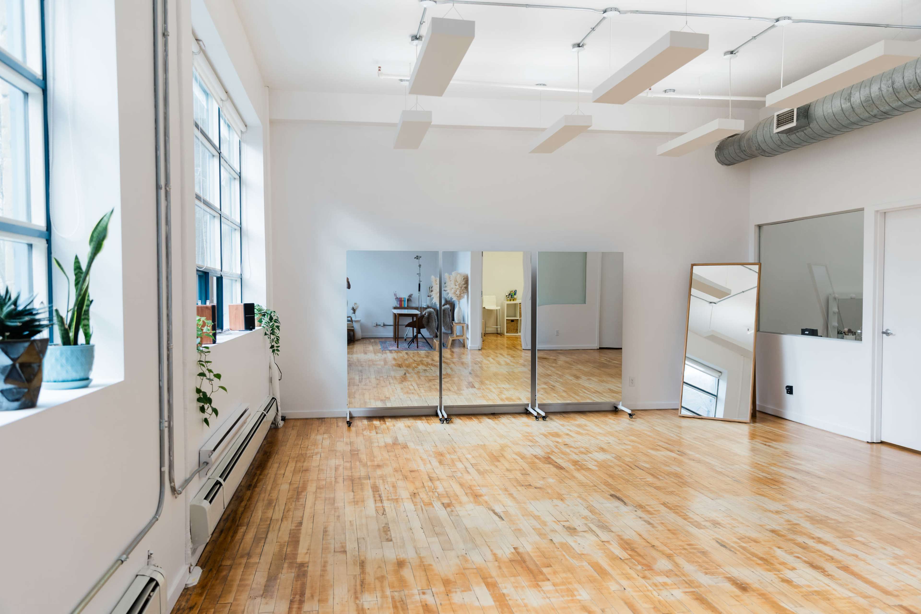 The image shows a spacious, bright room featuring large mirrors, wooden flooring, and windows with plants.