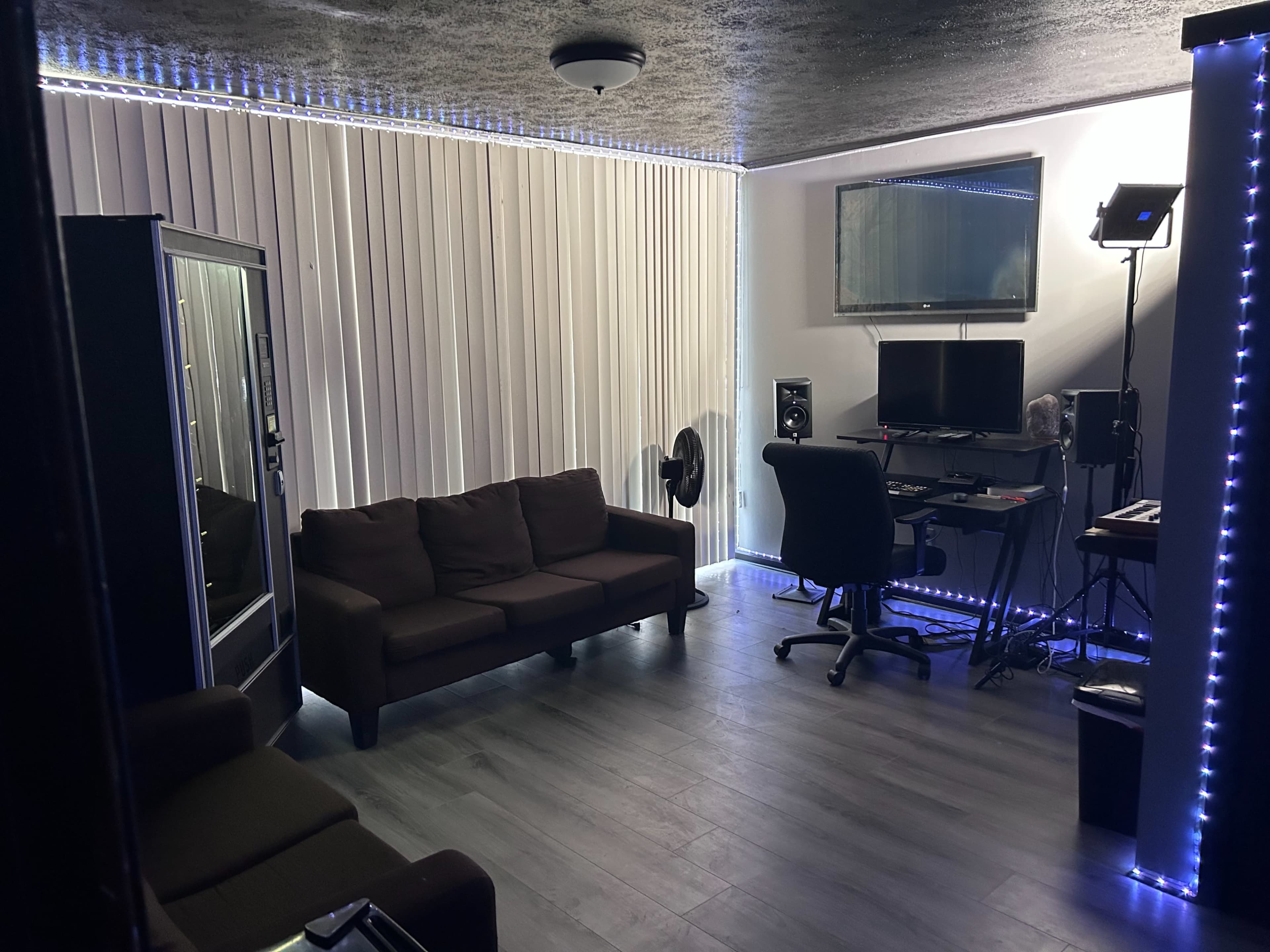 A small room features a brown couch, a desk with a computer and music equipment, a fan, and a tall mirrored cabinet, all illuminated by LED strip lights.
