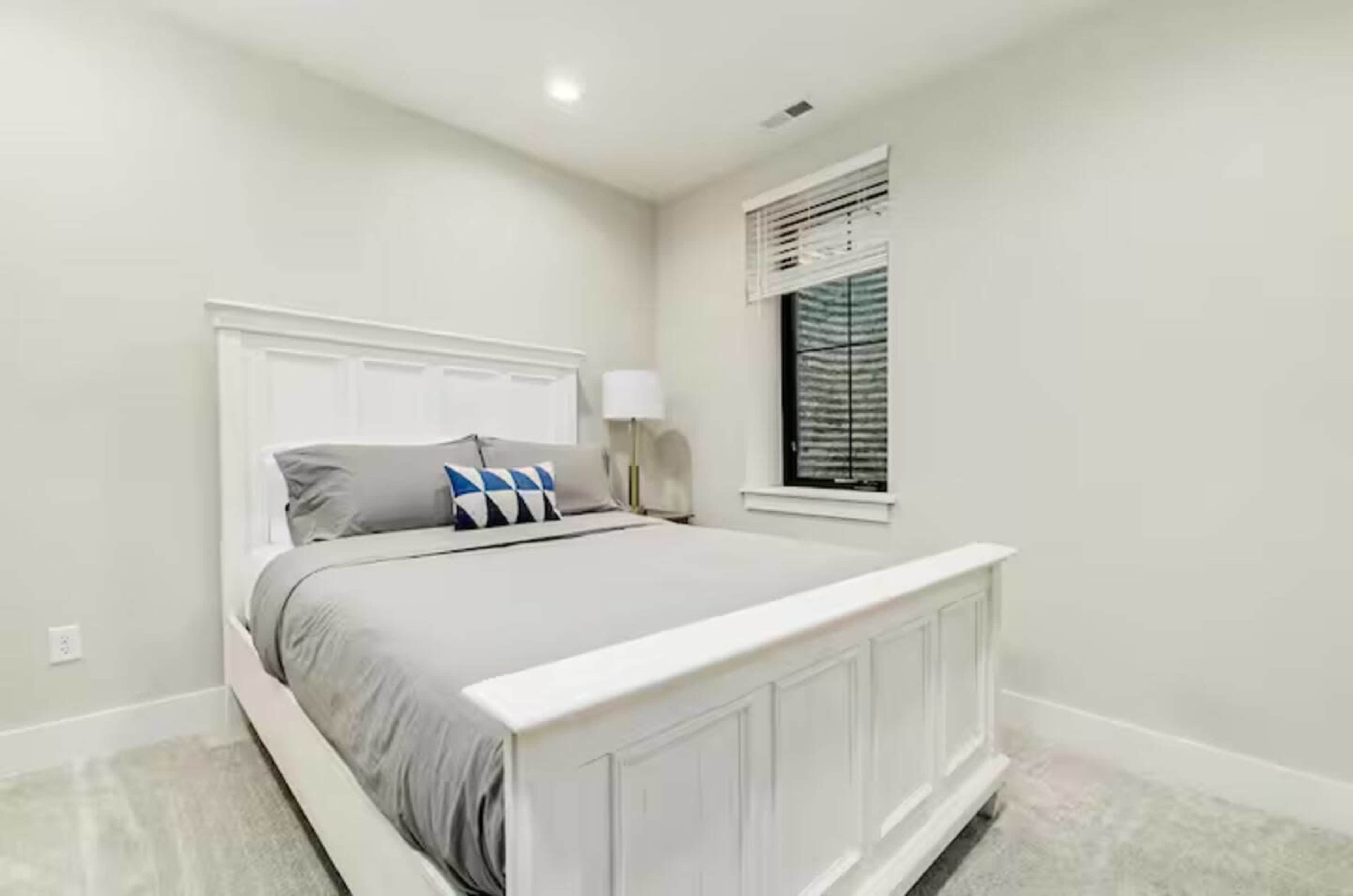The image shows a minimalist bedroom with a white bed frame, gray bedding, a decorative pillow, and a window with blinds.