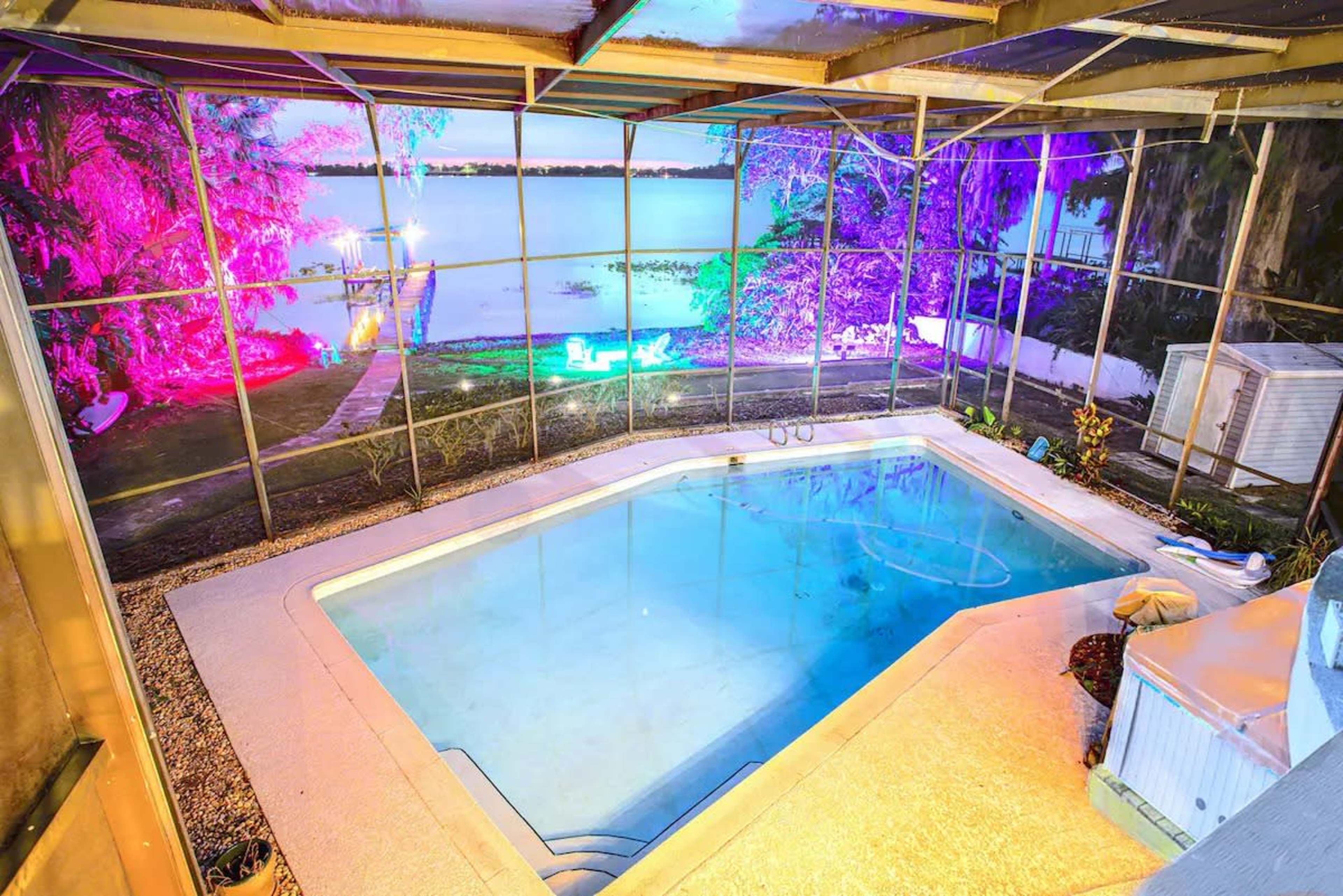 The image shows a pool enclosed by a screened patio, with colorful lights illuminating the surrounding area and a view of a lake in the background.