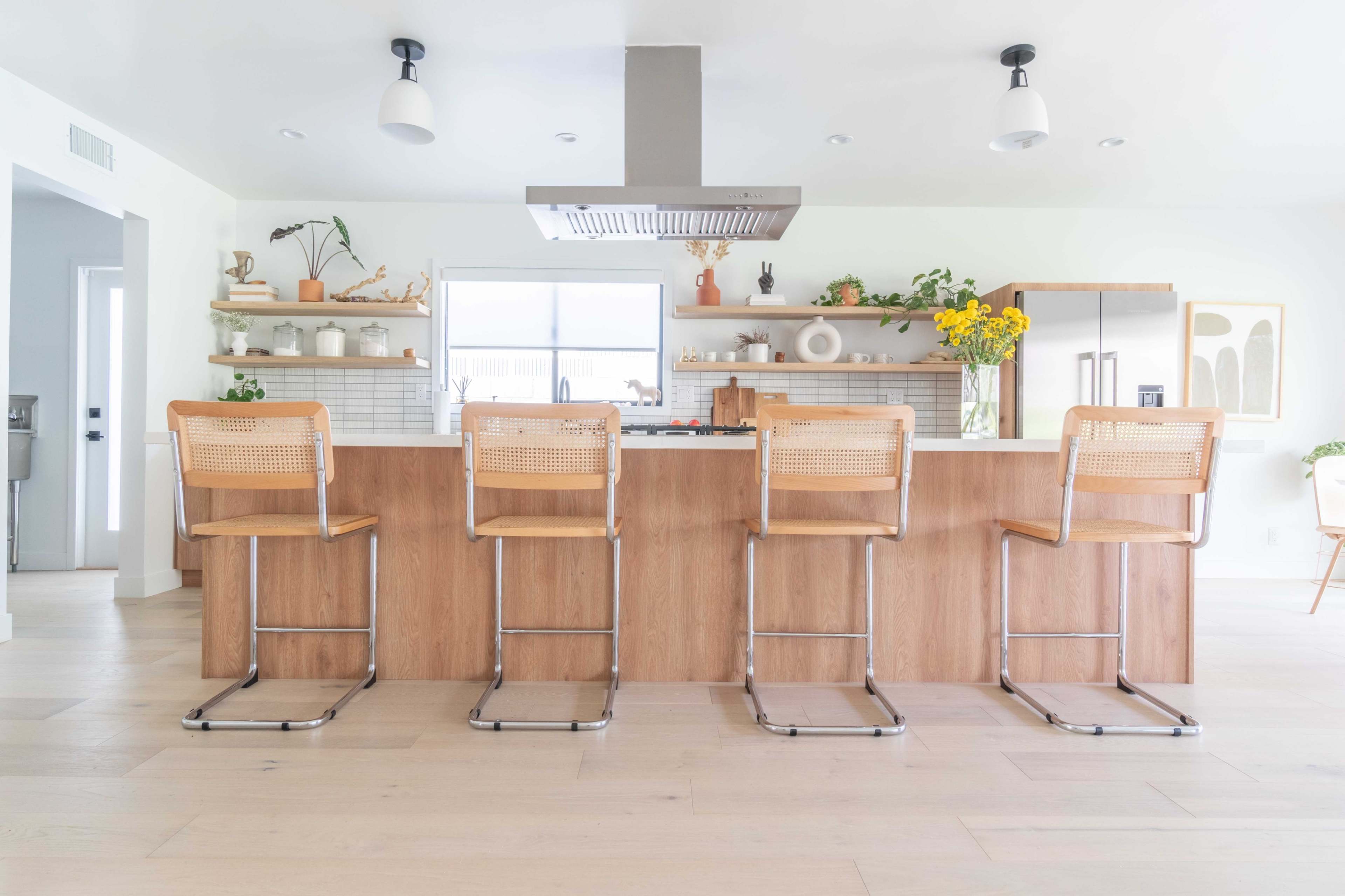 The image shows a modern kitchen with a long wooden island featuring four metal and wood bar stools, surrounded by light-colored walls and various decorative items on shelves.