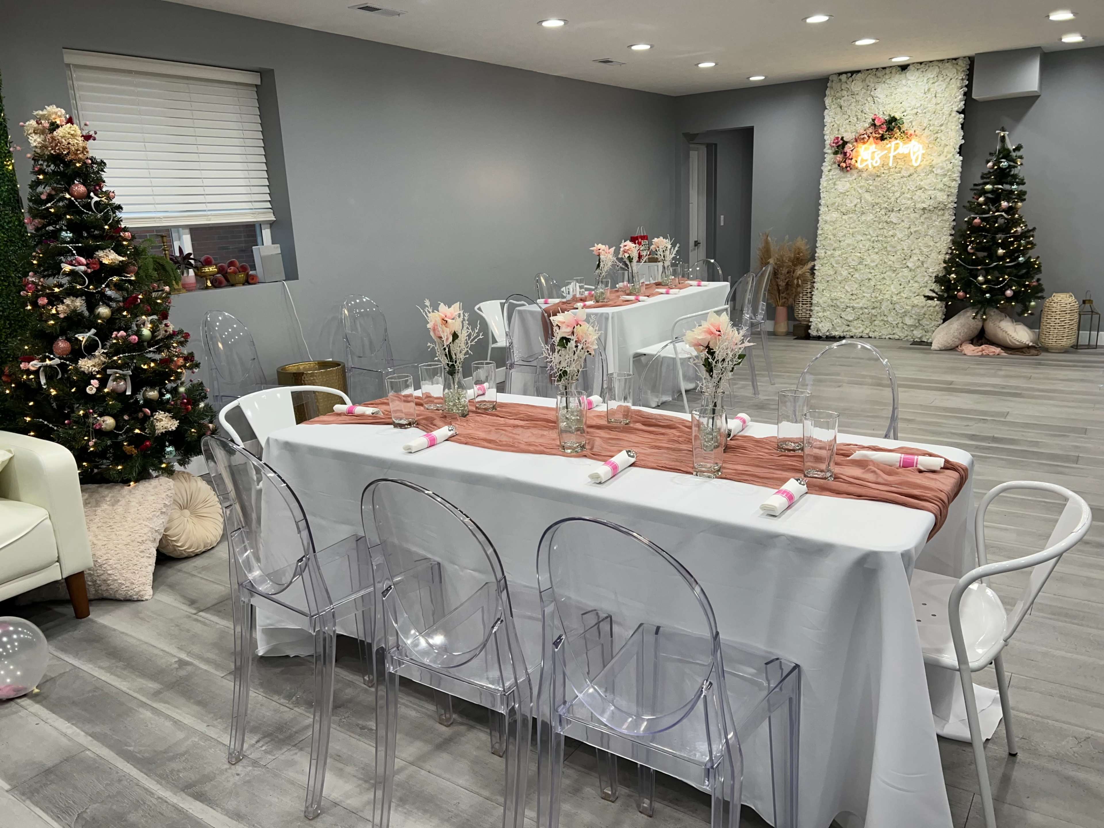 A decorated event space features two tables set for a gathering, adorned with floral centerpieces and surrounded by clear acrylic chairs, with Christmas trees in the background.