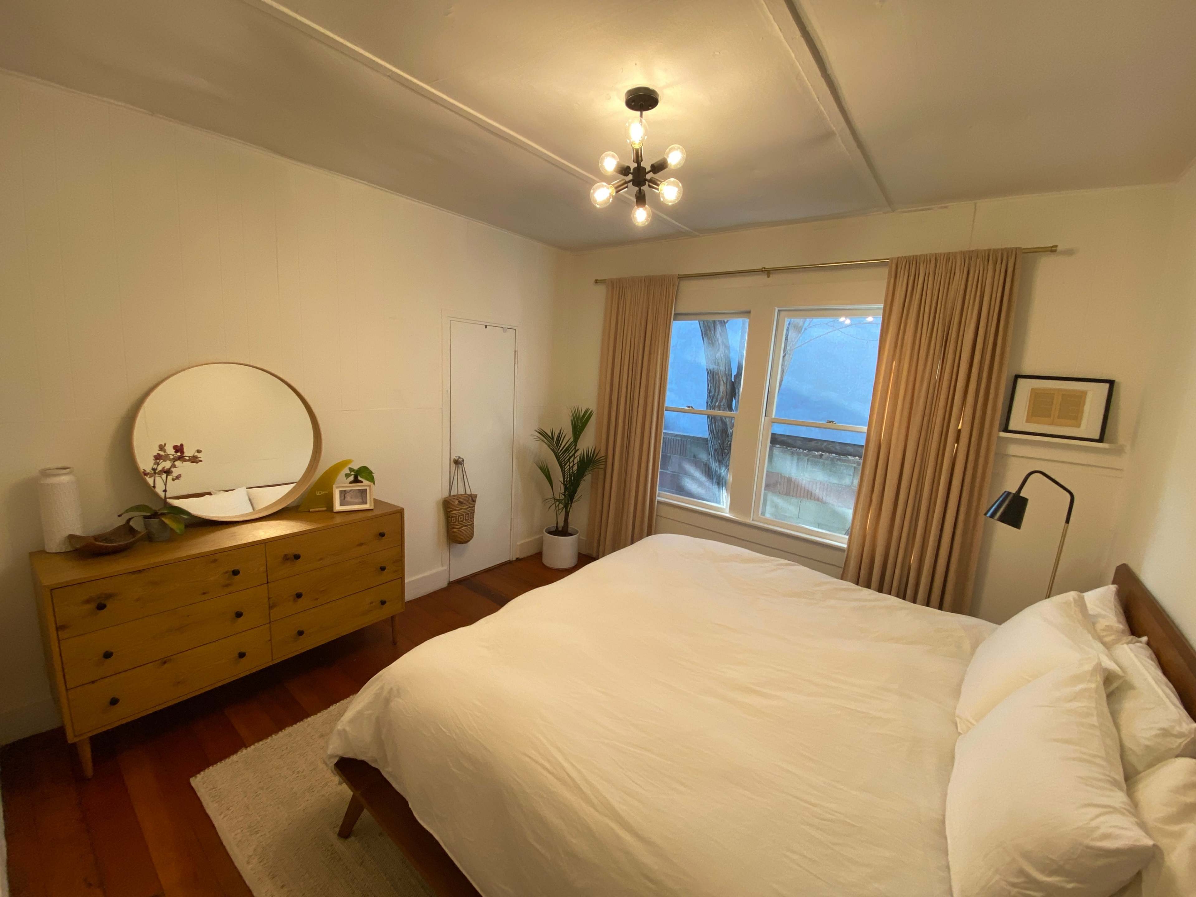 The image shows a bright bedroom with a bed, a wooden dresser, a circular mirror, window curtains, and a plant beside the window.