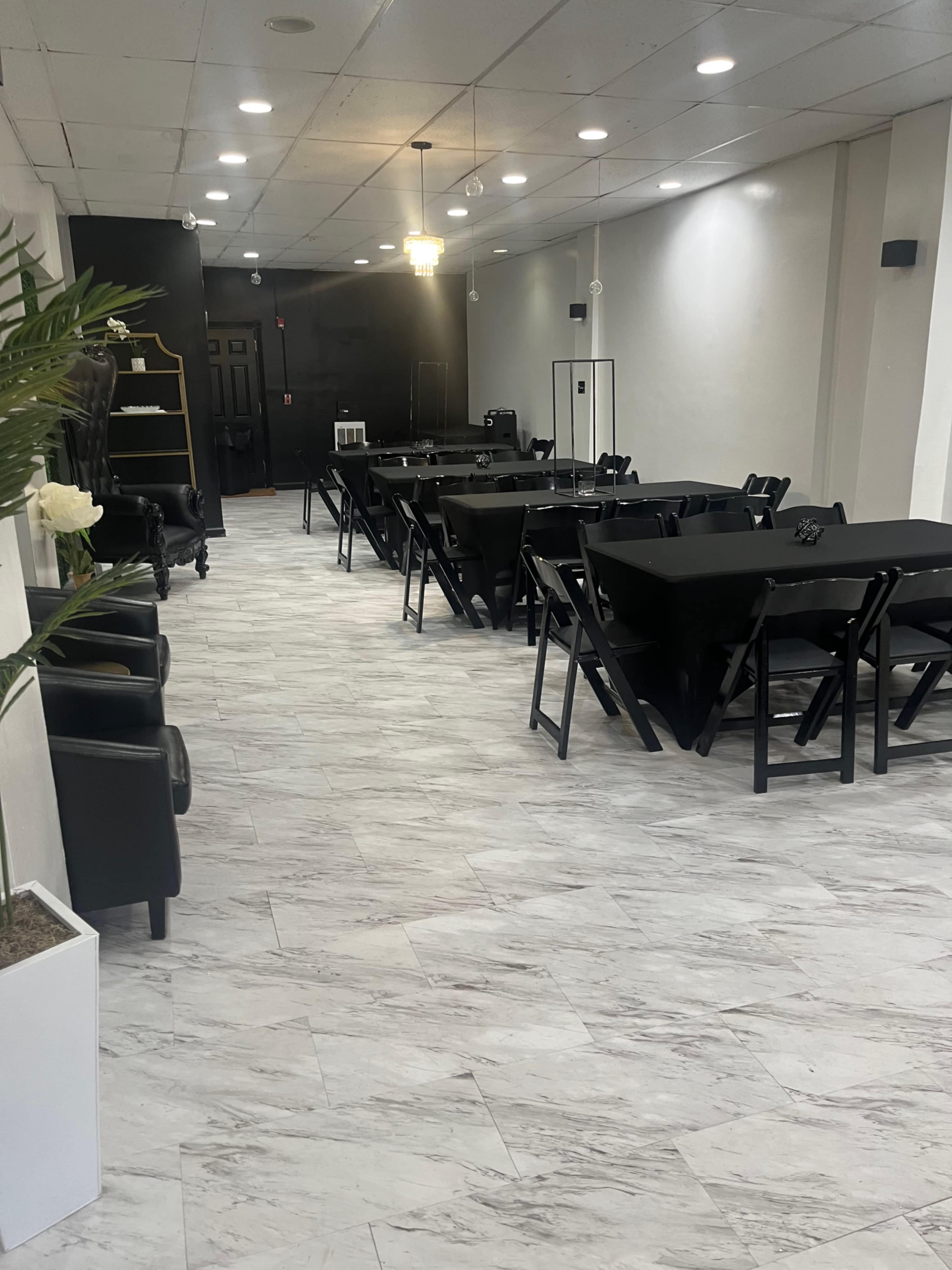 A spacious event room with several black folding tables and chairs arranged in rows, featuring light-colored tiles and a black accent wall.