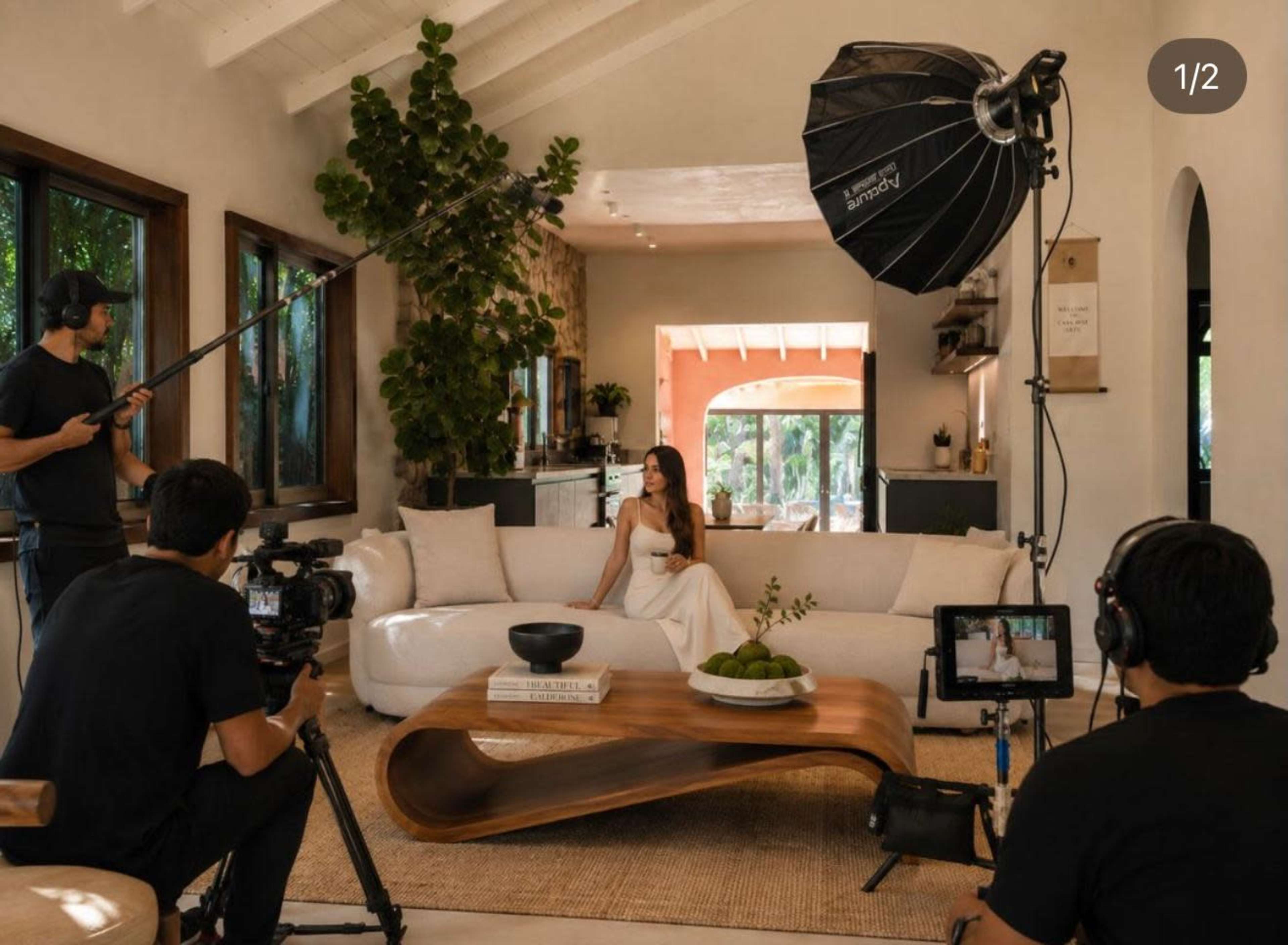 A woman sits on a white couch in a well-lit living room, while three people set up camera equipment and lighting for a photoshoot.