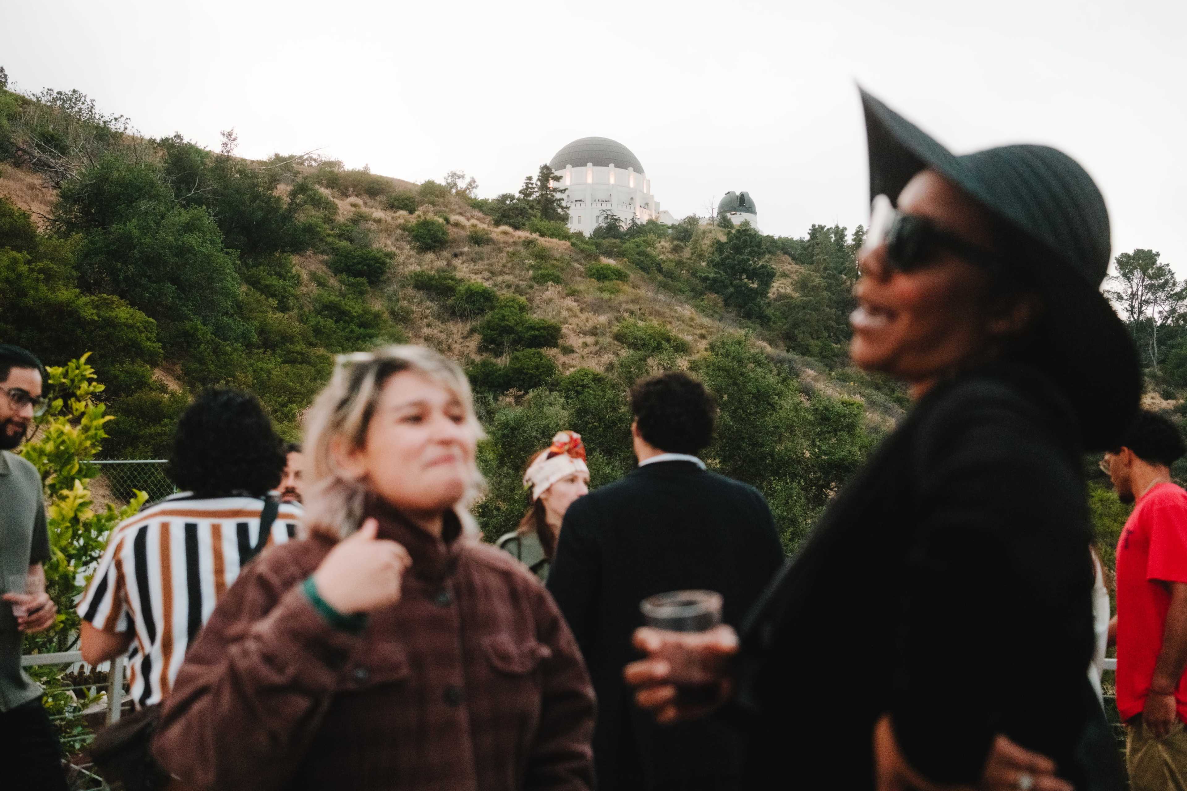 A group of people socializes outdoors with the Griffith Observatory visible on a hillside in the background.