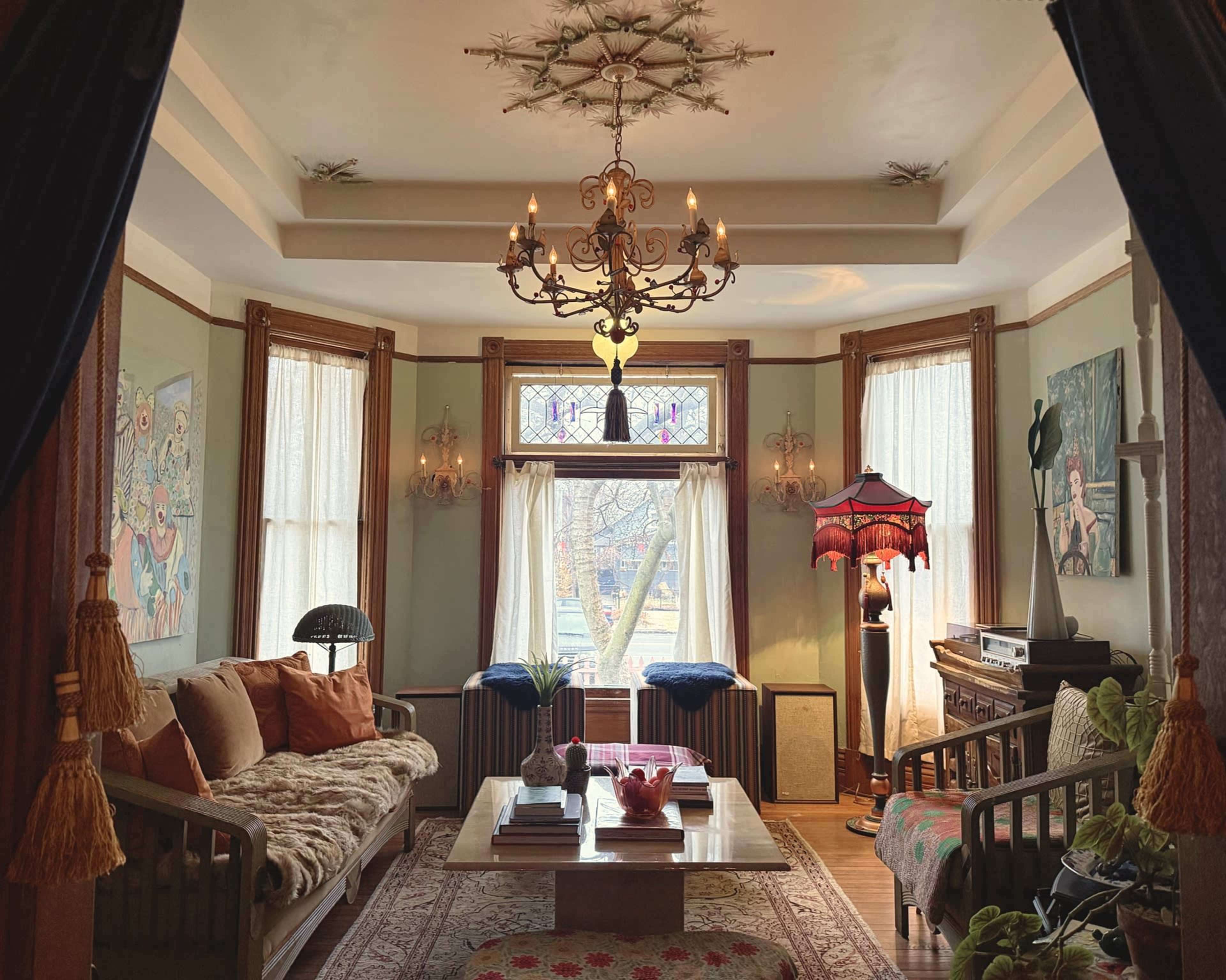 The room features a large sofa, a wooden coffee table, decorative lamps, and large windows draped with light curtains, creating a warm and inviting atmosphere.