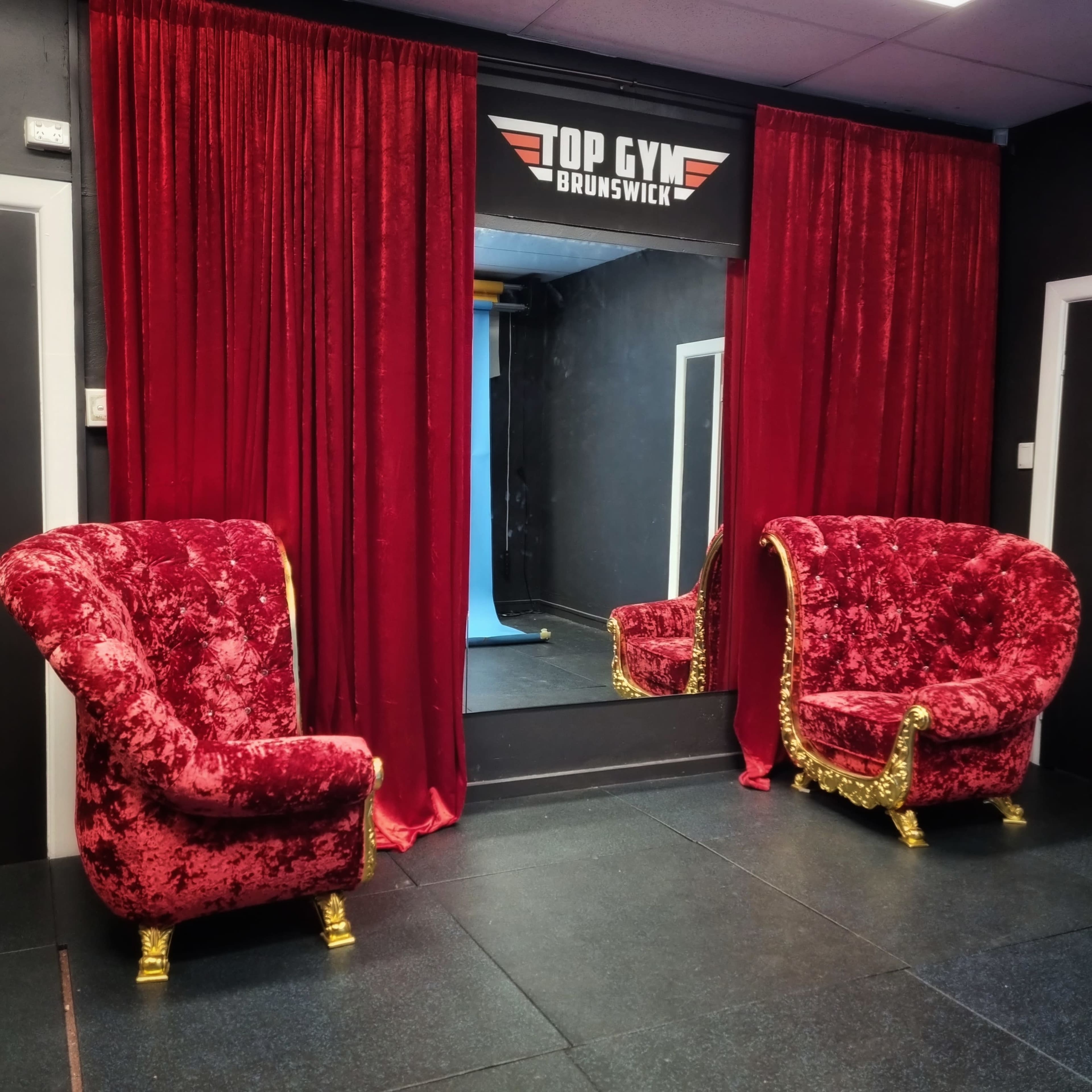 Two ornate red velvet chairs with gold accents facing a mirror, framed by red curtains in a gym setting.