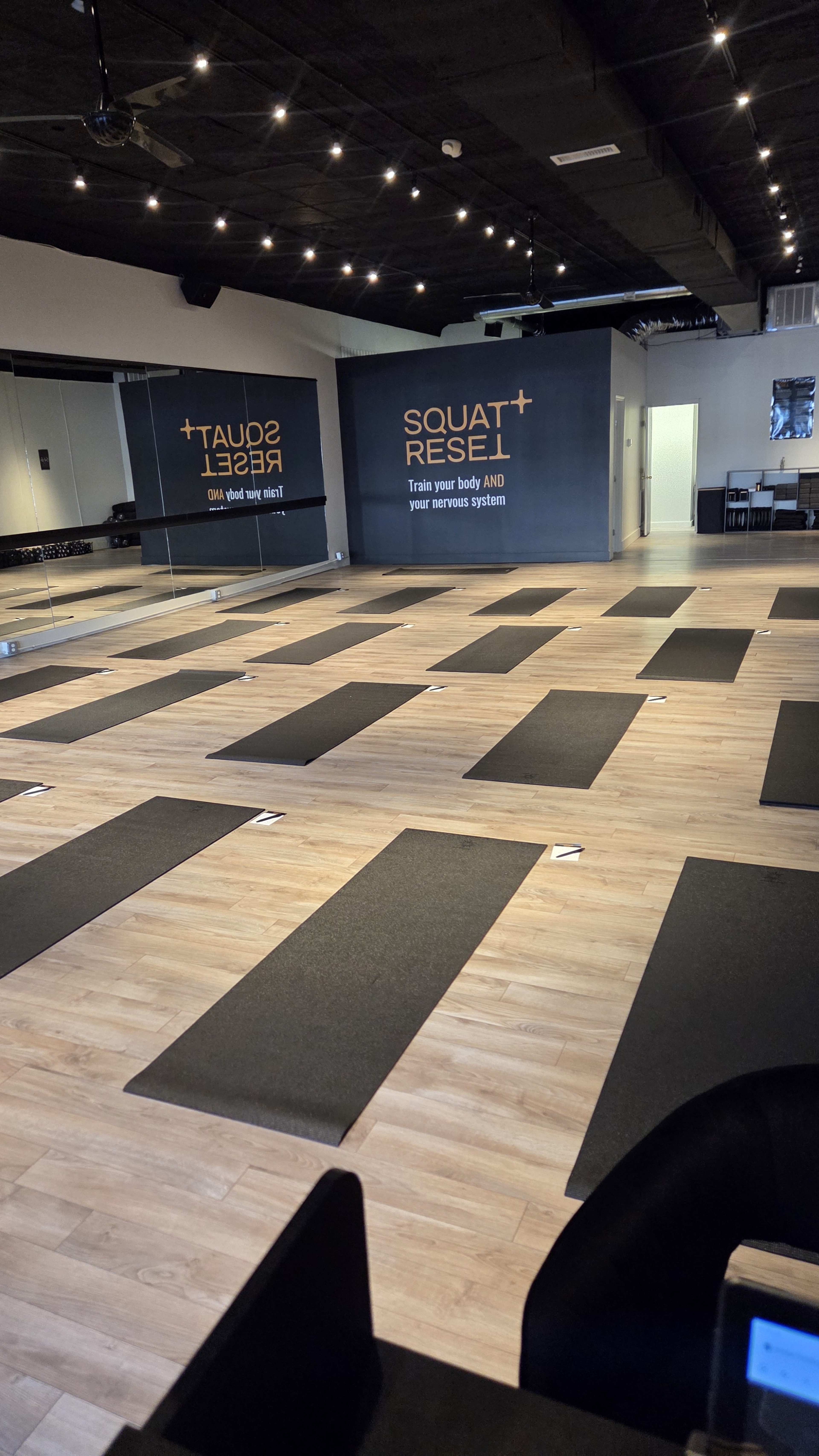 The image shows a spacious fitness studio with several yoga mats arranged on a wooden floor, and motivational text on the wall.