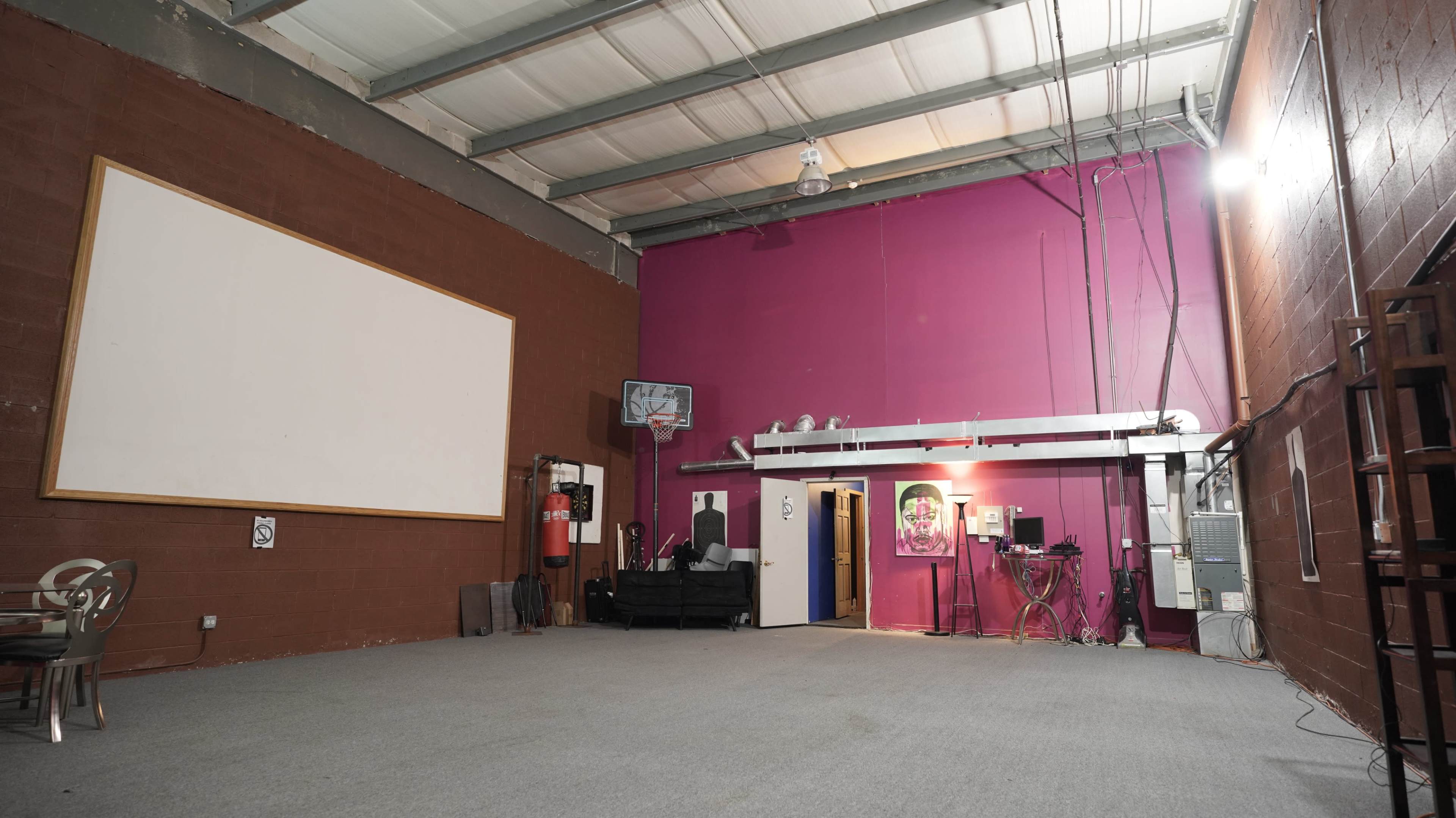An empty room with a high ceiling, a large blank wall on the left, and a vibrant pink wall on the right, featuring a door and various equipment along the back wall.